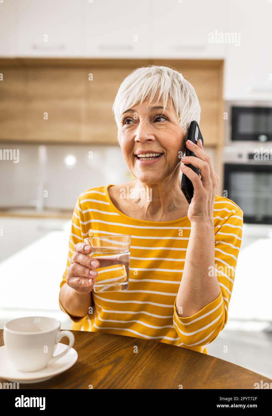 Pretty mature woman talking on mobile phone and holding glass of water at dining table at home Stock Photo