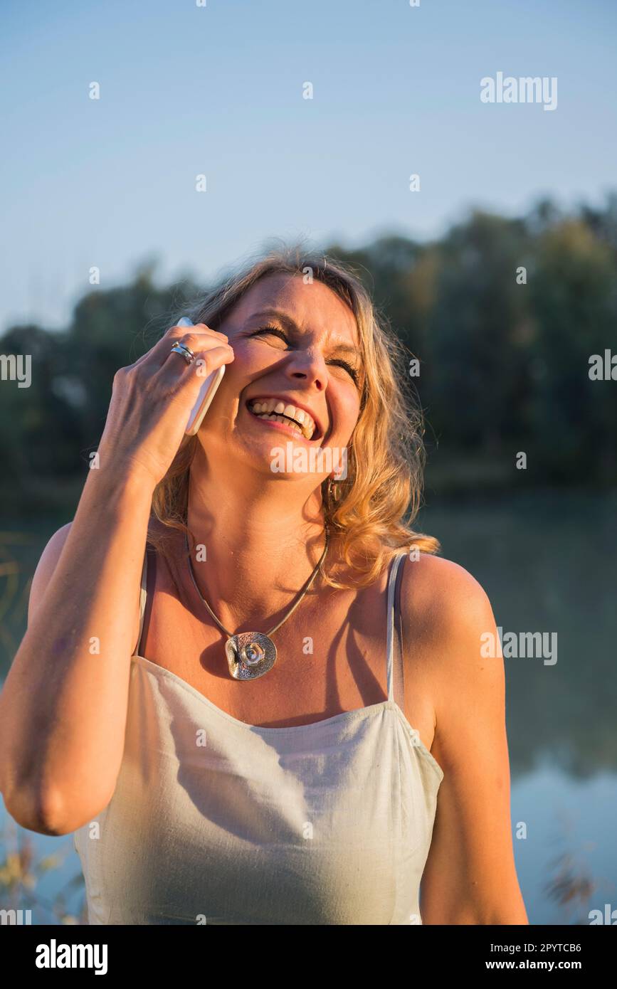 Cheerful woman laughing while talking on smart phone at riverbank, Bavaria, Germany Stock Photo