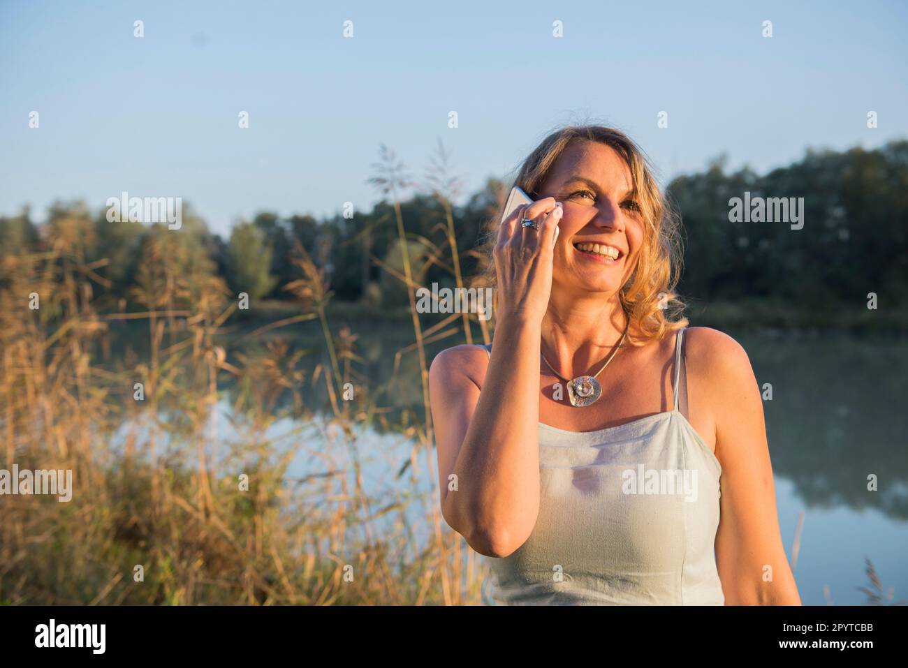 Cheerful woman smiling while talking on smart phone at riverbank, Bavaria, Germany Stock Photo