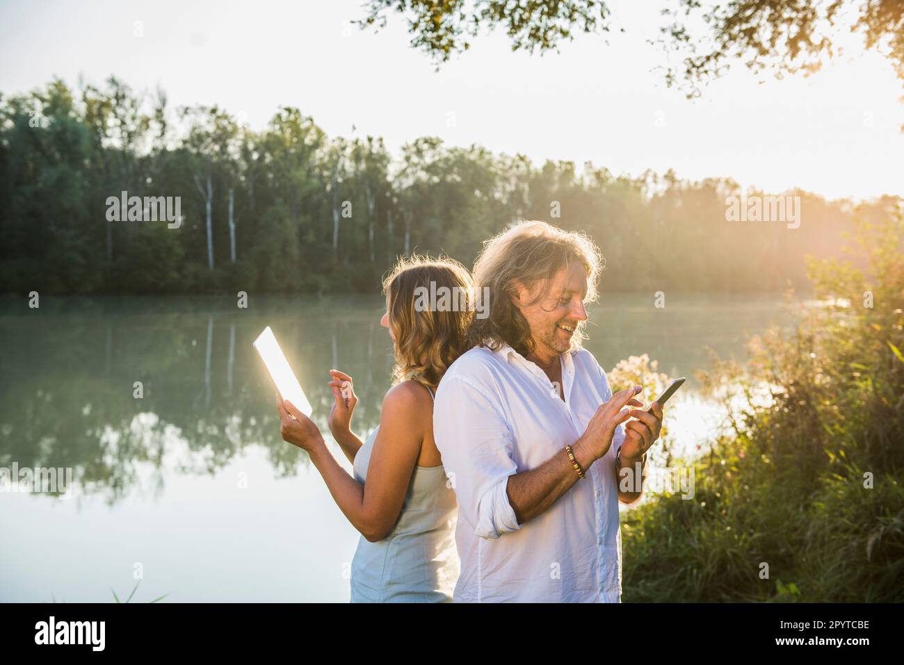 Couple at a river using smart phone and digital tablet, back to back Stock Photo