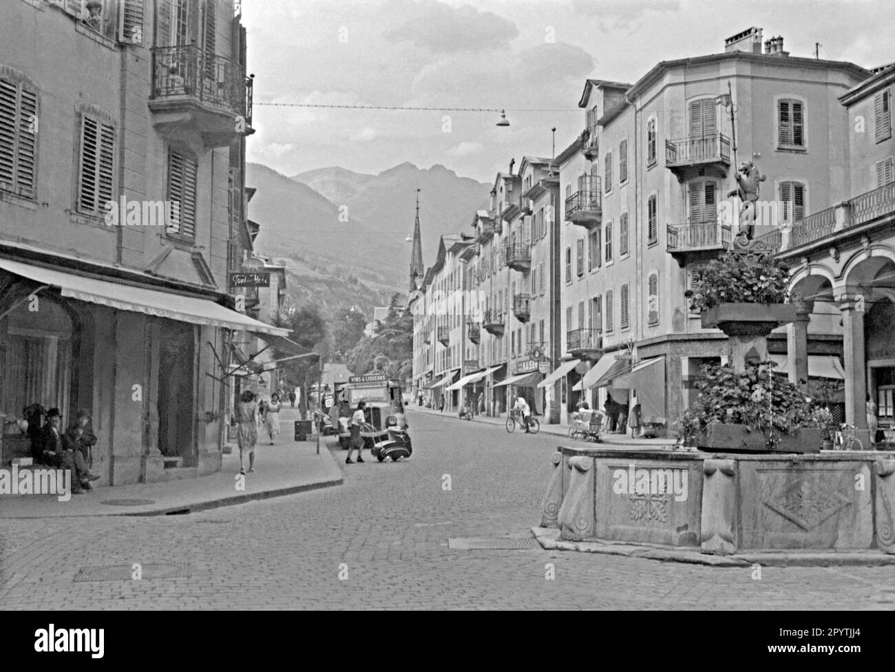 A street scene featuring a fountain in the Old Town, Chur, Graubunder, Switzerland in 1949. The water feature forms a ‘roundabout’ at the convergence of several streets in the town centre. It is capped by a sculpture of a bear holding a standard. Shops are open and shoppers are out and about. A drinks delivery truck is parked in the road. This is from an old amateur black and white 35mm negative – a vintage post-war photograph. Stock Photo