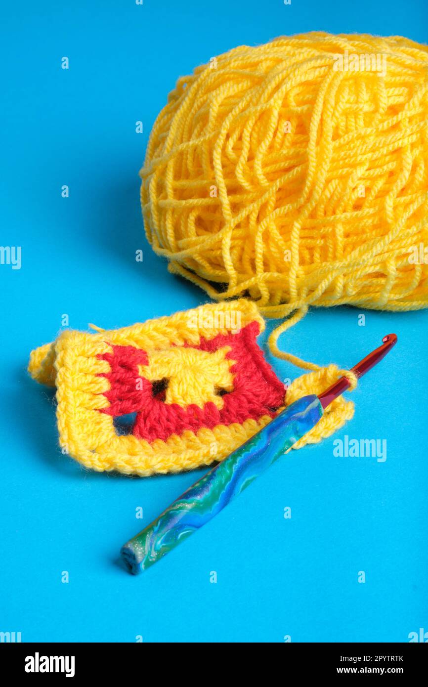 Crochet Granny Square being made with yarn and hook, yellow and red on kingfisher blue background. Stock Photo