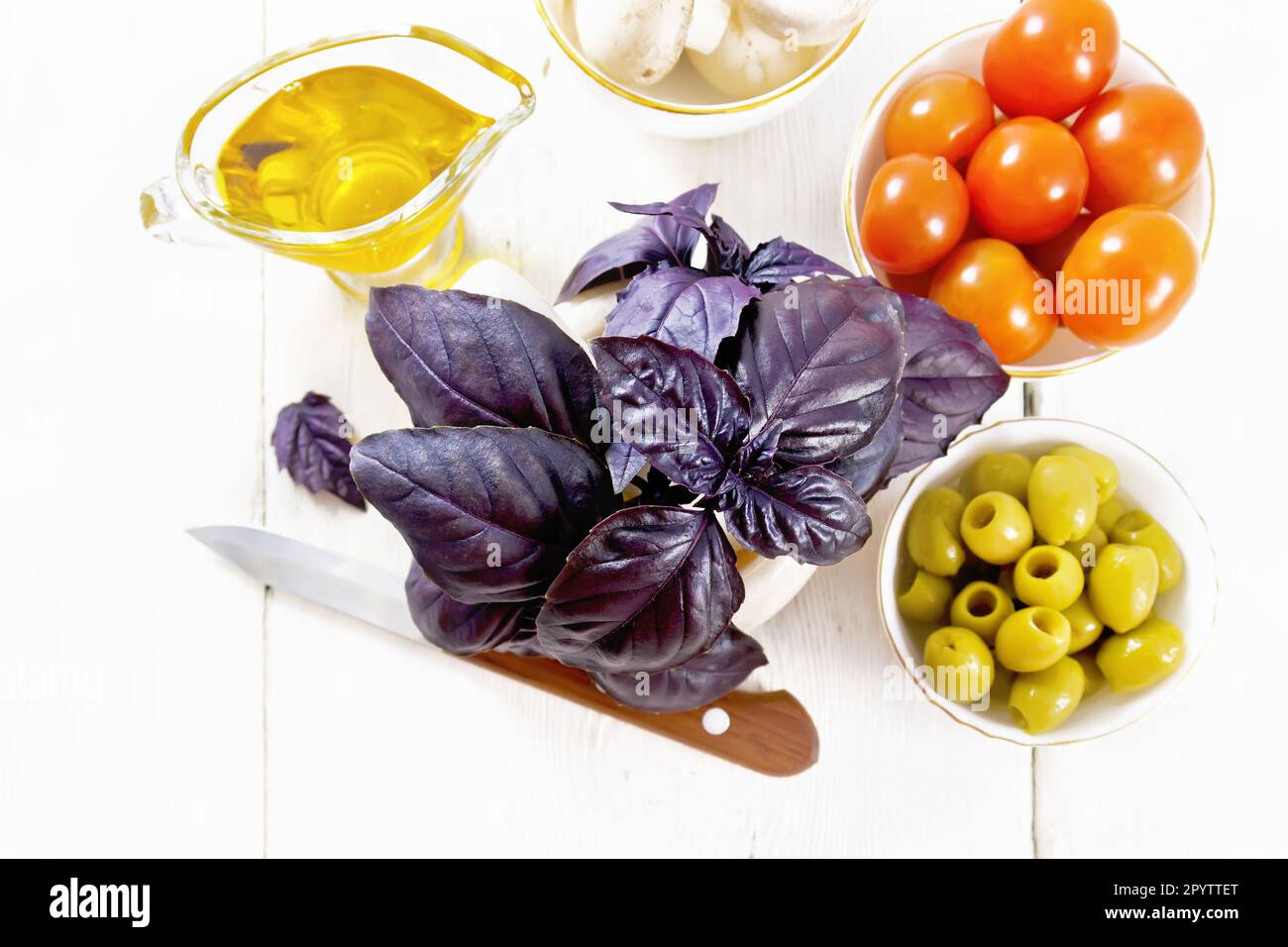 Fresh purple basil in a mortar, olives, tomatoes and champignons in bowls, vegetable oil in gravy boat and a knife on light wooden board background fr Stock Photo
