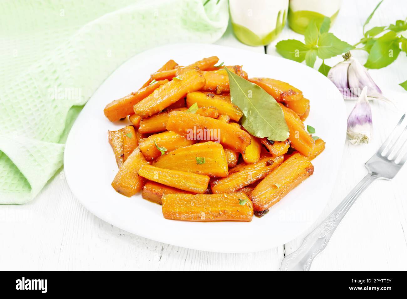 Carrots fried with garlic, bay leaf, spices and green onions in a plate, basil and napkin on white wooden board background Stock Photo