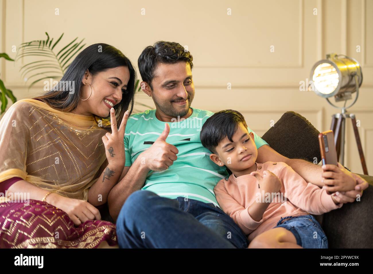 Happy realxed family with kid taking selfie on mobile phone while sitting on sofa at home - concepts of technology, togetherness and family bonding. Stock Photo