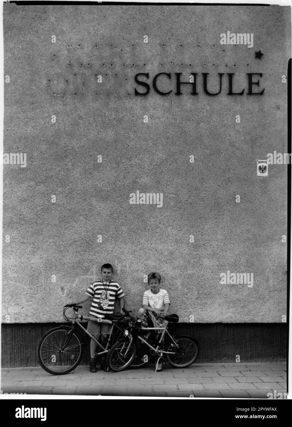 ' East Germany, Strausberg. After the fall of the Wall. Change of name of the former ''Karl-Marx Oberschule'' to ''Schule''. Wall with lettering, in front of it 2 boys with their bicycles. Street scene, black and white. Photo, 1995.' Stock Photo