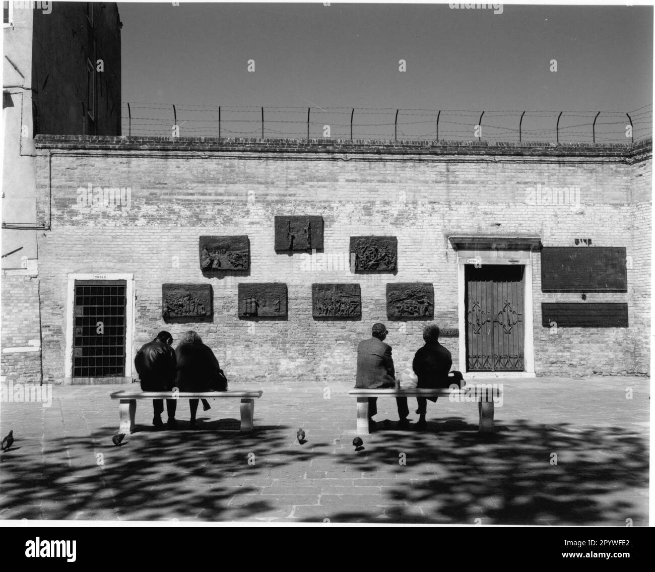 Venice, Venezia (Italy), Ghetto (Jewish Quarter), Campo del Ghetto Nuovo, Holocaust memorial - Monumento all’olocausto (seven bronze reliefs, unveiled 1980, sculptor: Arbit Blatas, 1908–1999). Visitors sit at the memorial. Street scene, black and white. Photo, 1997. Stock Photo