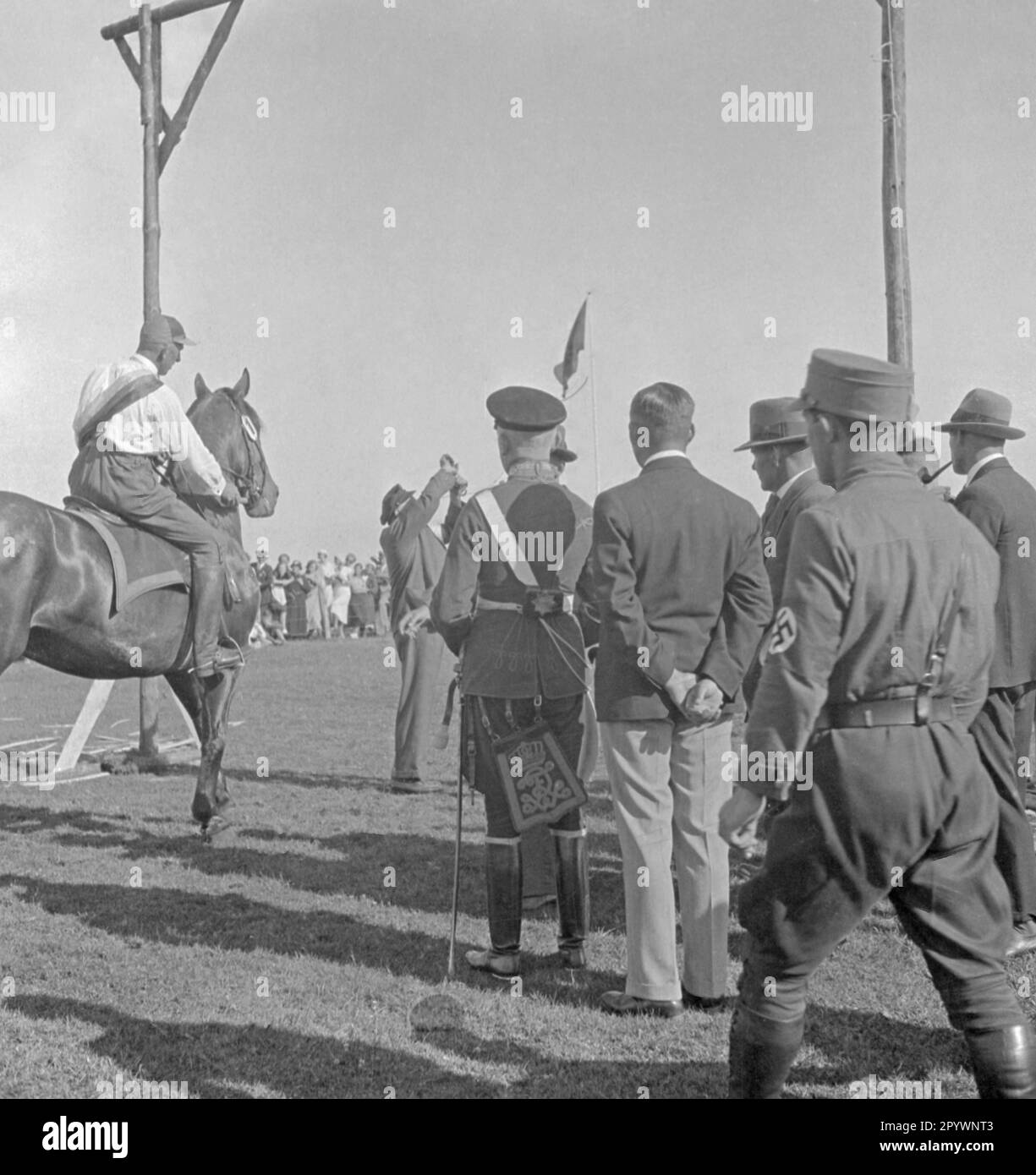 Rider participates in ring riding in Warnemünde. In ring riding, a lance is put through a ring hanging from the scaffold. Spectators stand next to the scaffold. A man wears a uniform of the Prussian army. Right in the picture is a SA man. Stock Photo