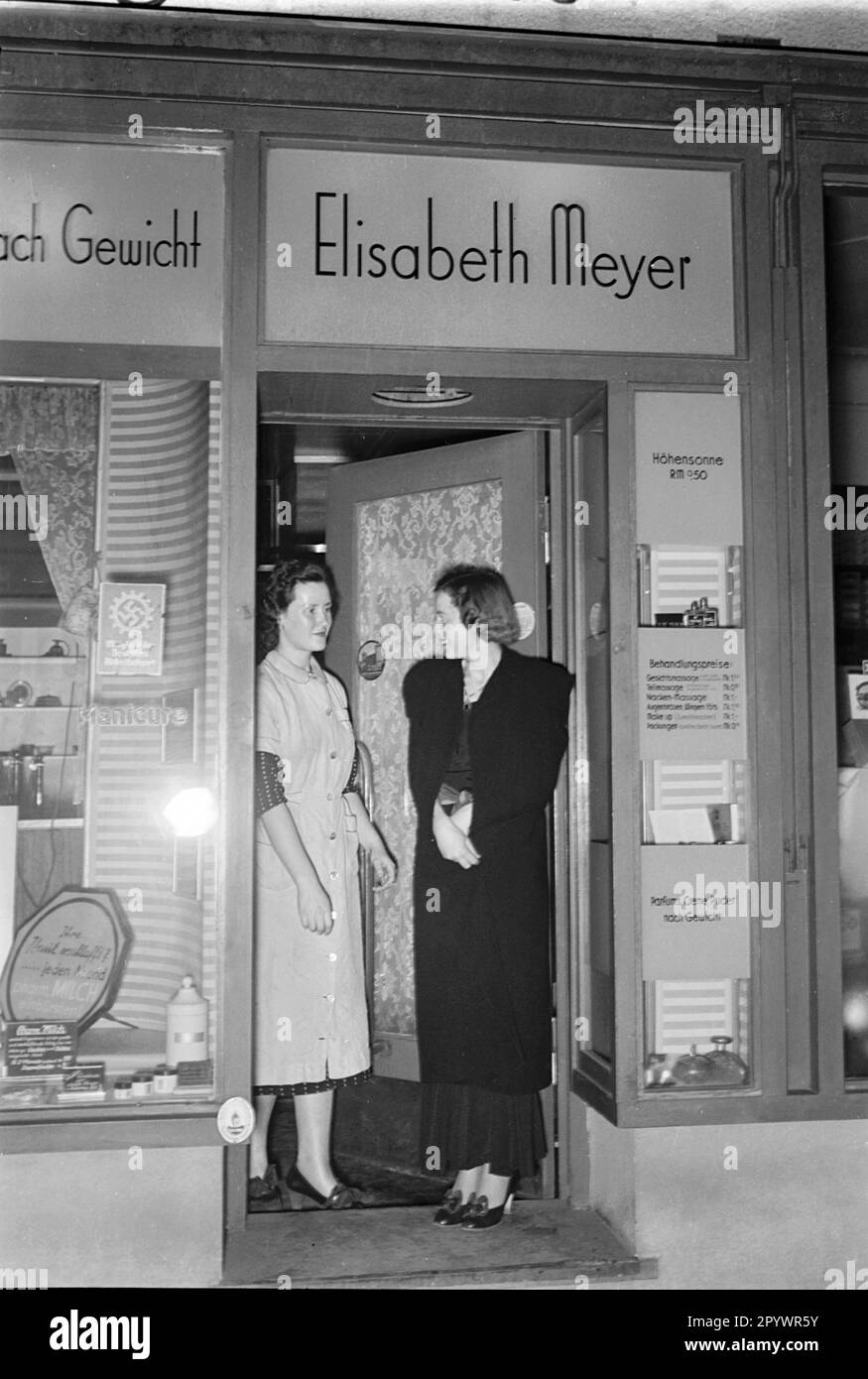 A customer leaves the beauty salon of Elisabeth Meyer. In the shop window on the left is a sign of the German Labor Front (DAF), on the right an advertisement for a treatment with sunlamp. Stock Photo