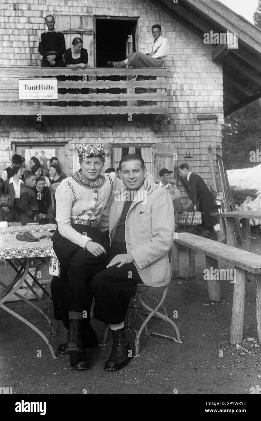 Couple sitting in front of a hut in an Austrian ski resort.  In the background are further skiers. Undated picture, probably from 1938. Stock Photo