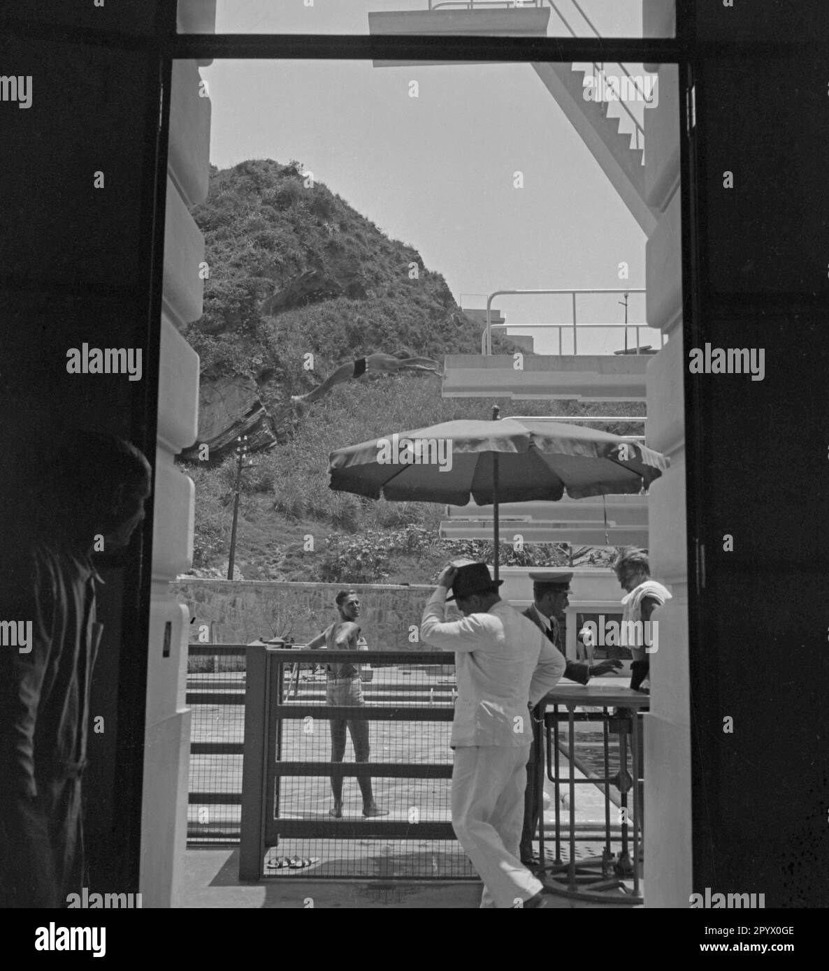 Look through a door onto the entrance area of the swimming pool. In the middle stands a boy at the edge of the pool, while in front of him another boy jumps from the diving tower. On the right are two men at a turnstile, presumably the entrance to the swimming pool. Stock Photo