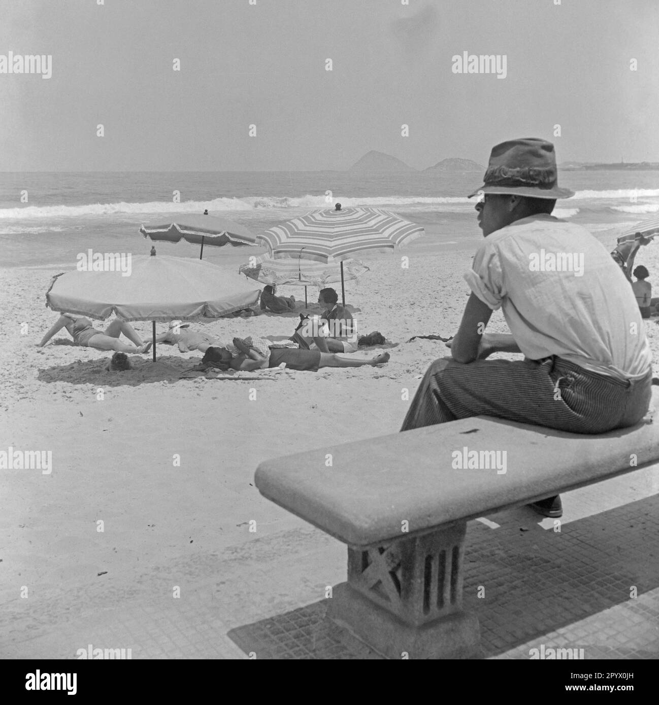 A Brazilian boy sits on a bench at the beach (presumably Avenida Atlantica / Copacabana). In front of him is a group of tourists under umbrellas or while sunbathing. Stock Photo