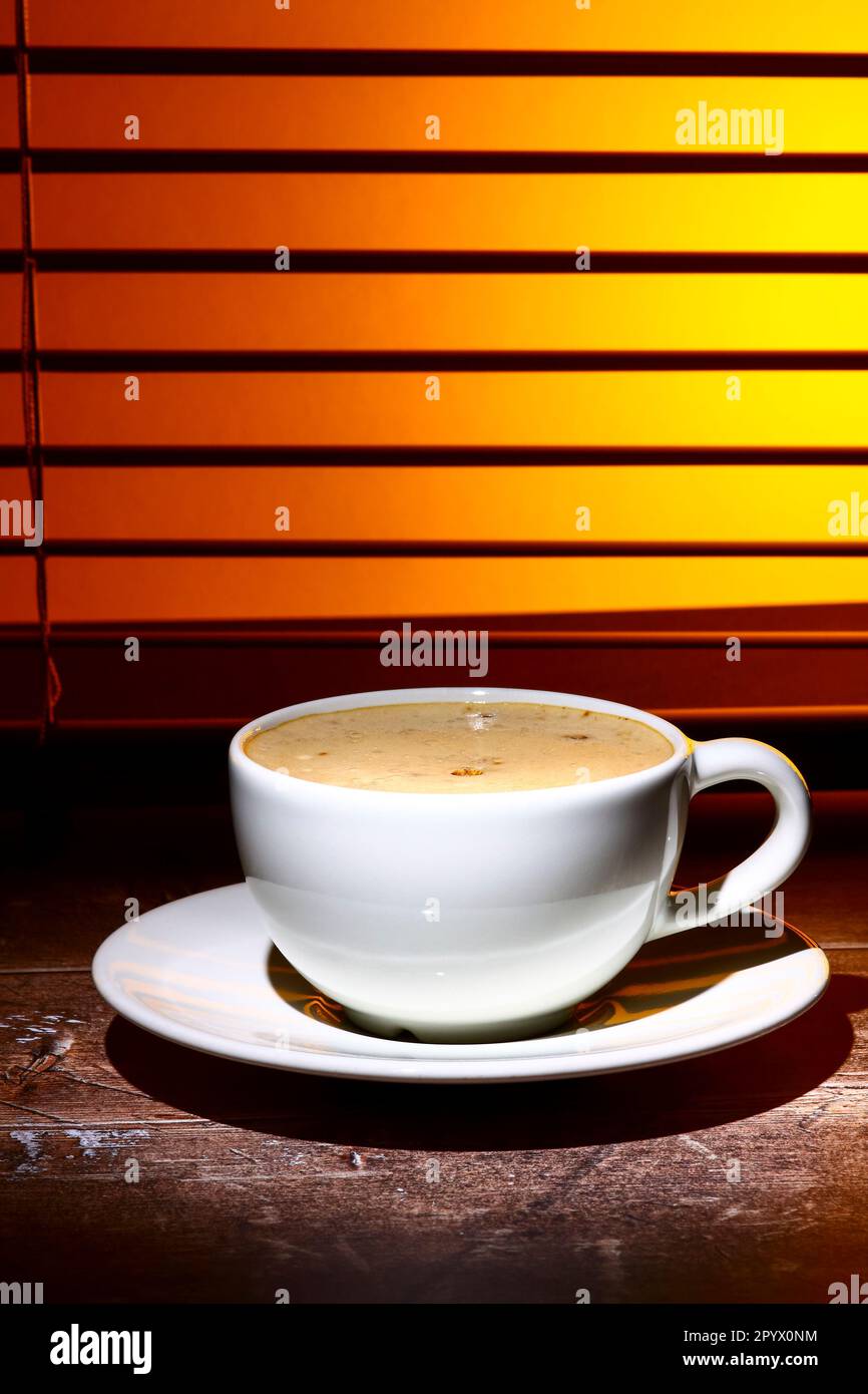 coffee cup filled with frothy coffee set against a window blind Stock Photo