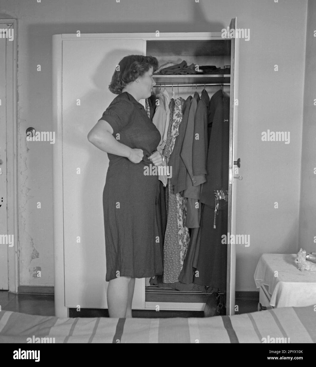 A woman stands in front of an open wardrobe in a bedroom and examines herself in the mirror of the closet door, while touches her waist with her hands. Stock Photo