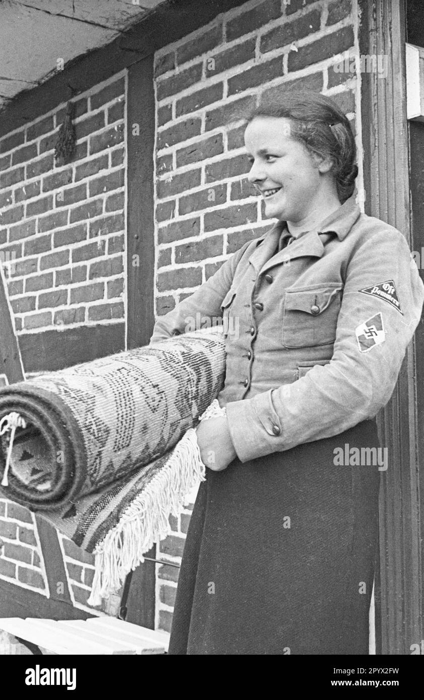 A young woman takes a finished carpet out of the house. She works and lives in a small artisanal business in Lubmin as part of the Landfrauenjahr (compulsory year of service of women in the countryside). On the upper arm of the BDM uniform is the patch Gau Pommern. Stock Photo