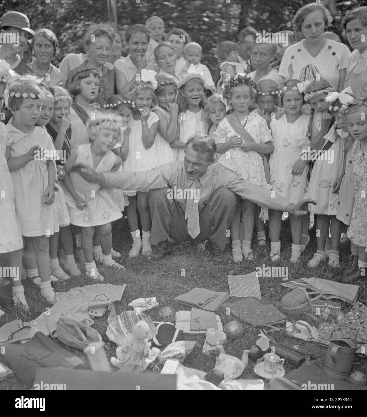 A man spreads his arms in front of many little girls dressed in white clothes, in front of him are various everyday objects on the ground, such as hats, cloths, jugs, dolls, baskets, etc. Stock Photo