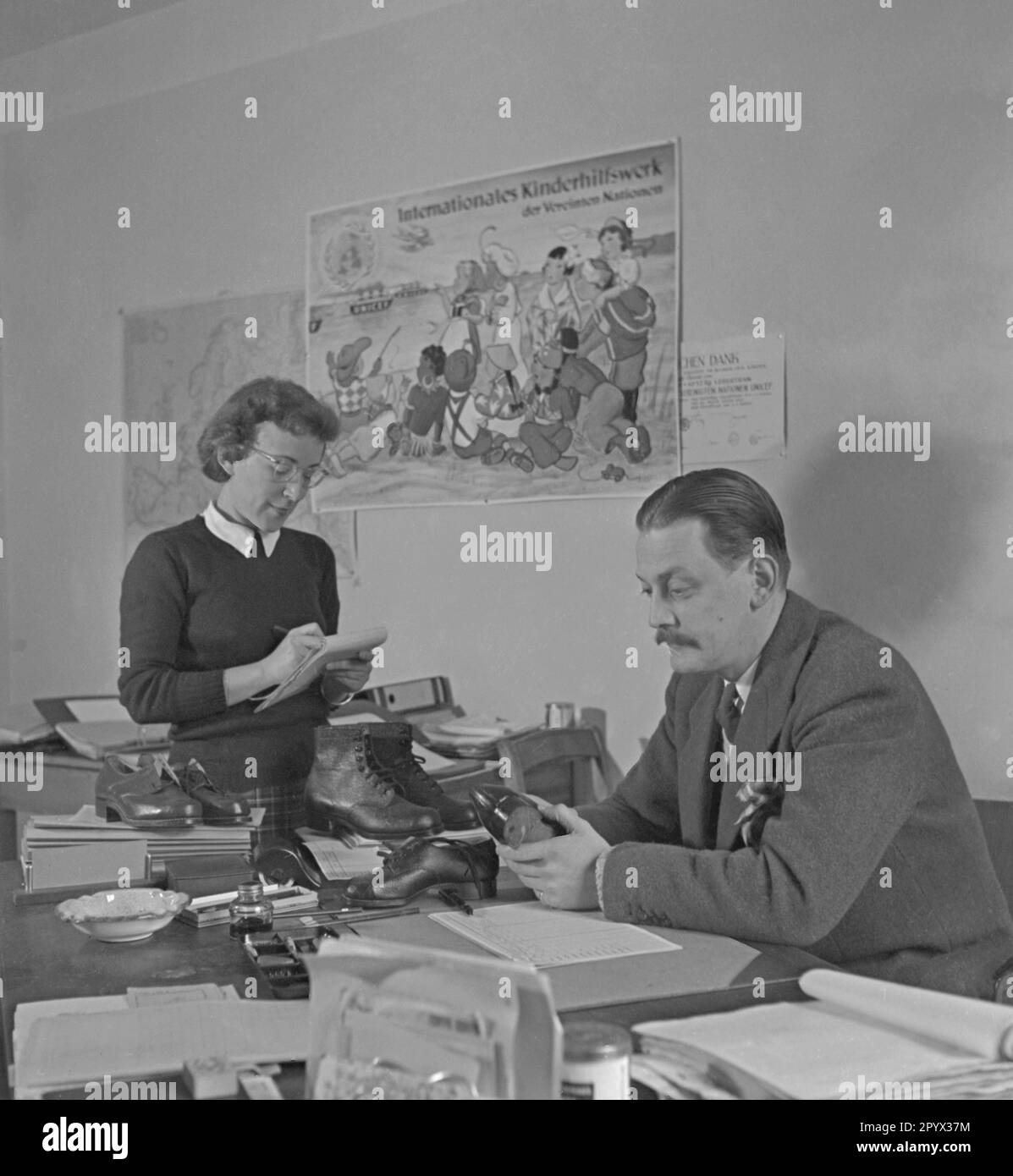 At an office United Nations Children's Fund, an employee inspects shoes made by seamstresses. On the left a secretary. On the wall hangs a poster of UNICEF. Stock Photo