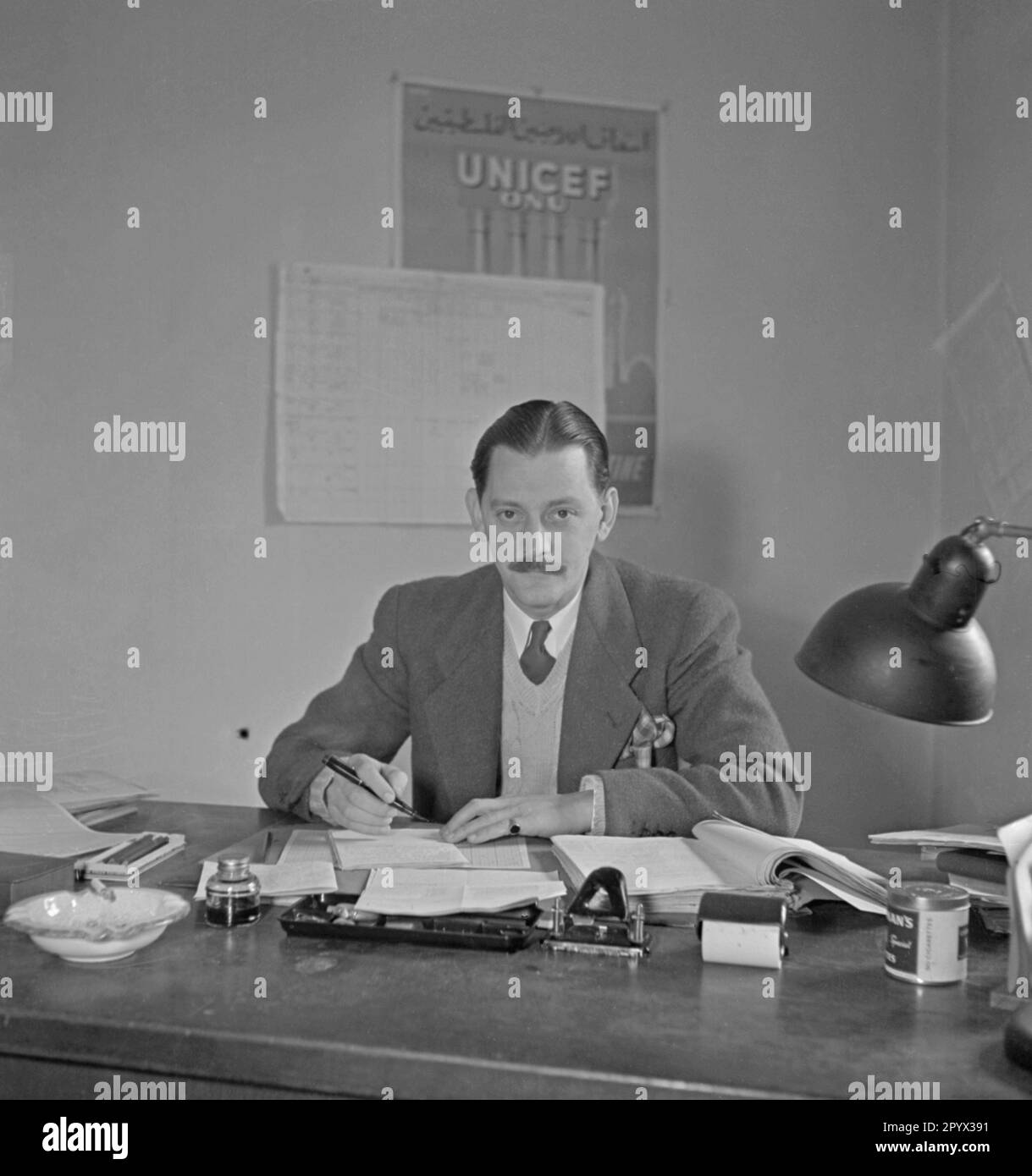 An employee of the Berlin Children's Fund of UNICEF at his desk. Undated photo. On the wall hangs a poster of UNICEF and the ONU (UNO) with Arabic characters. Left, an ashtray with cigarette and an inkwell. Stock Photo