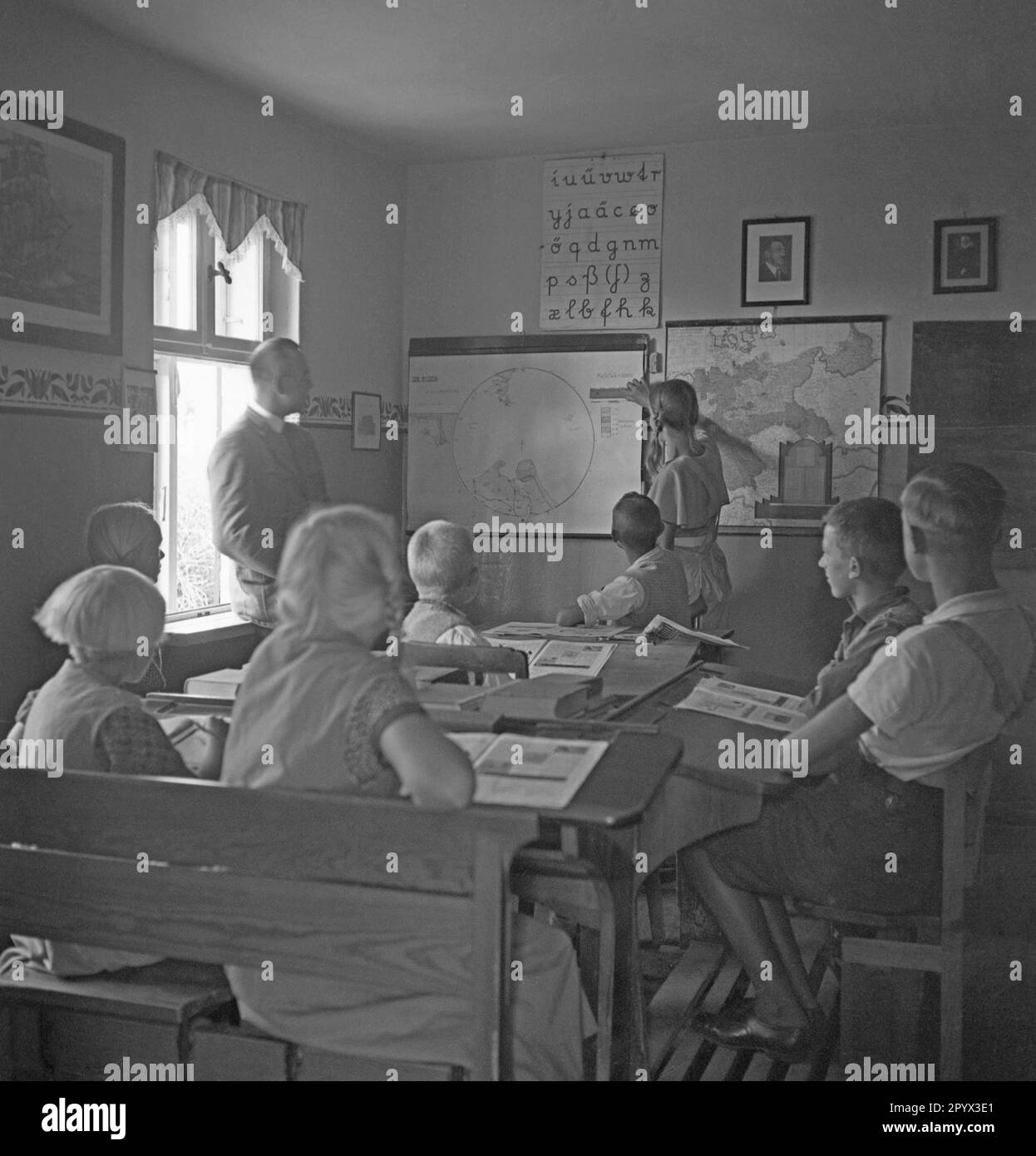 In a school on the Baltic Sea island of Ruden a teacher teaches students geography and local history. A student measures the scale with a ruler on a map showing the Baltic Sea coast and the island of Ruden and Usedom. Above a map of Germany hangs a portrait of Adolf Hitler. Stock Photo