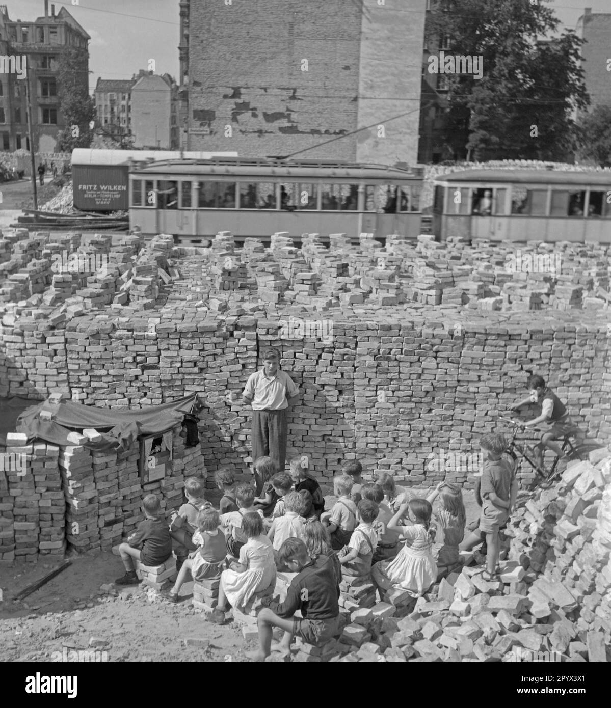 Undated photo of a group of children watching a puppet show among piles of bricks remnant from the ruins. Bricks serve as curtain of the puppet theater. In the background, more brick stacks, a passing street car and partly destroyed houses by bombing. Stock Photo