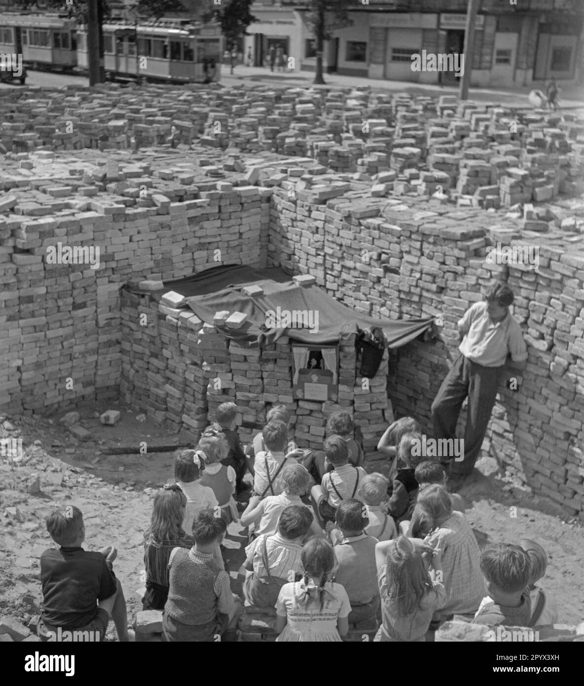 Undated photo of a group of children watching a puppet show among piles of bricks remnant from the ruins. Bricks serve as curtain of the puppet theater. To the right of the stage, the puppeteer. In the background, more brick stacks and a passing tram. Stock Photo