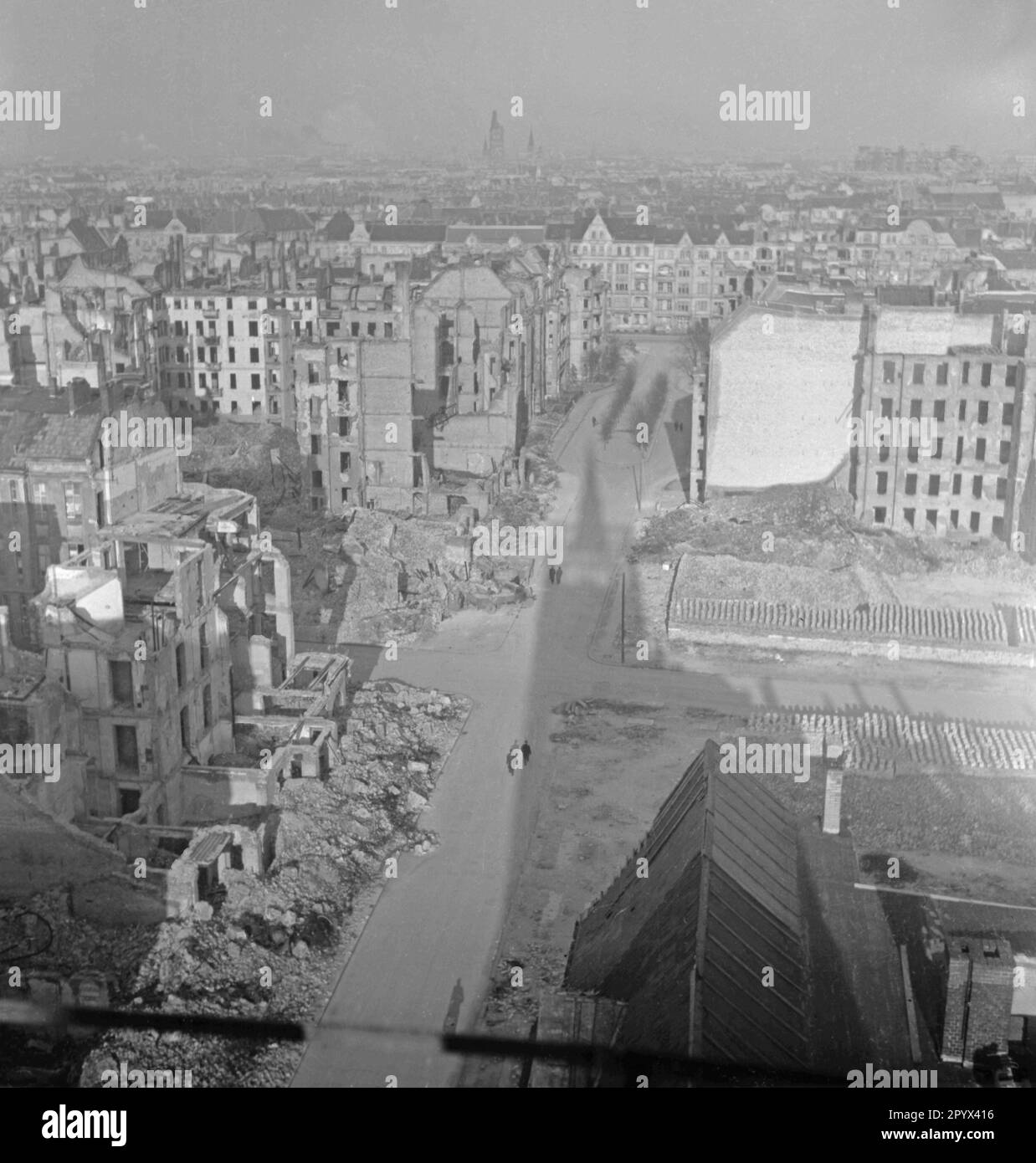 Undated photo of a partially destroyed almost deserted street in Berlin, presumably West Berlin, 1950. To the left and right of the road, partially destroyed houses, as well as house frameworks. To the right of the road, beginning of decluttering. Here, stacks of bricks for reconstruction. In the background, undamaged houses. On the horizon in the fog, the ruined tower of the Kaiser Wilhelm Memorial Church. Stock Photo