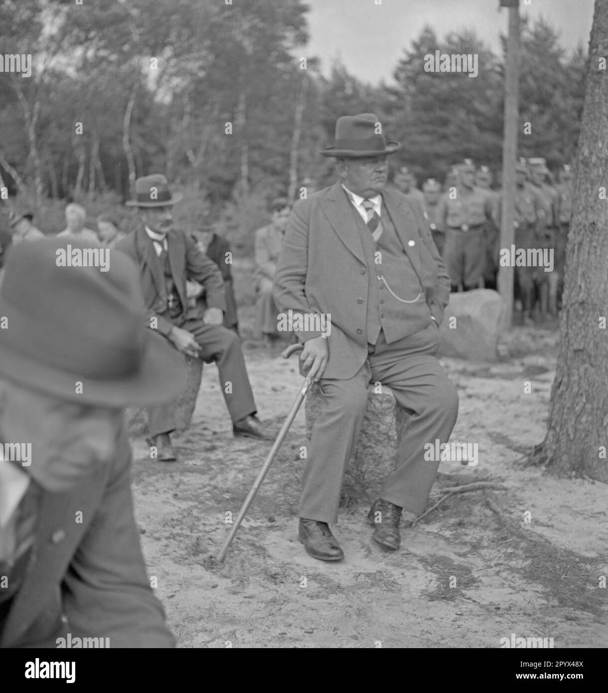 Photo of three local farmers' representatives (in a suit with a walking stick, sitting on a glacial erratic) of the neighbor communities at the inauguration of the Landtagsplatz in Hoesseringen at Suderburg in the Lueneburg Heath on June 28, 1936. In the background, a crew of the SA and spectators. Stock Photo