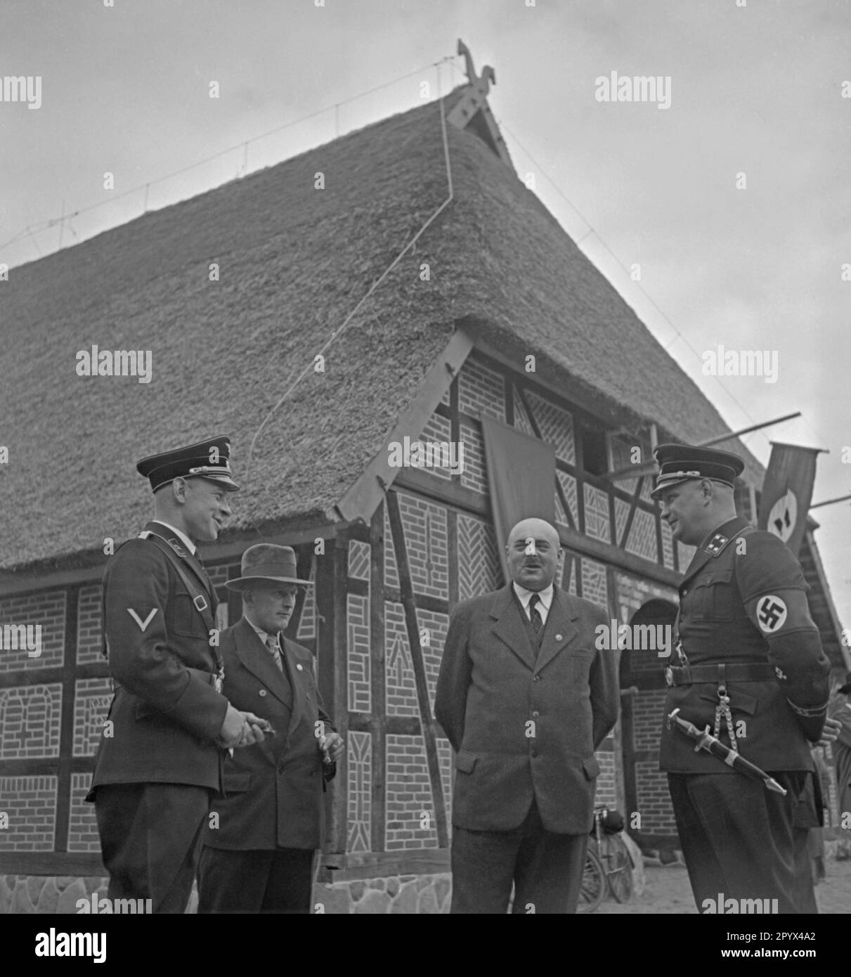 Photo of a group of Nazi farmer functionaries and SS men in uniform (on the left, an SS attack-man, on the right, an SS Sturmbannfuehrer) in front of the Agricultural Museum at the Landtagsplatz in Hoesseringen at Suderburg in the Lueneburg Heath. The occasion was the inauguration of the Landtagsplatz by the National Socialists on 28 June, 1936. At the entrance of the farmer's house (Low German aisled house with thatched roof and gable ornamentation, horseheads) meet farmers' representatives (some in SA uniforms) and bystanders. Swastika flags are hanging on the gable. Stock Photo