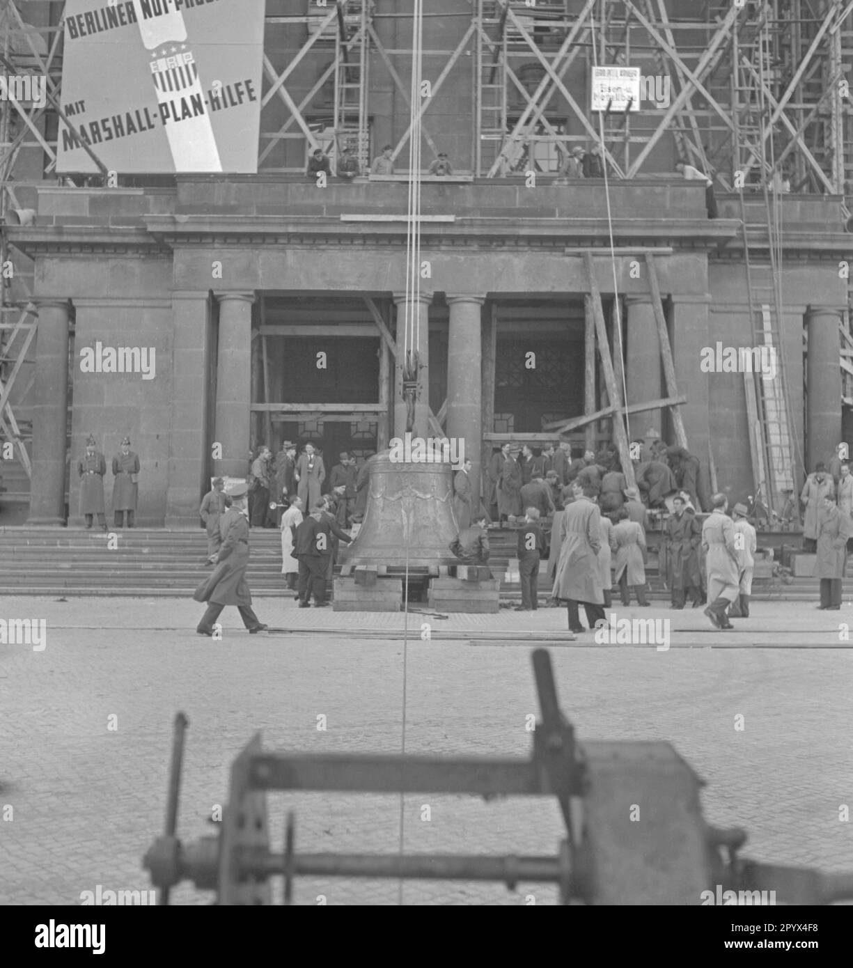 Photo of the Freedom Bell on Rathausplatz being pulled up by a crane from a wooden frame shortly before its installation in the tower of the governing mayor's seat, Ernst Reuter (1948-1953) in Berlin on October 21, 1950. Next to it, U.S. soldiers, policemen, passers-by and journalists. Behind, the columns of the main doorway and the tower scaffolding with a poster of the American occupation forces (inscription: Emergency Programme of Berlin with European Recovery Program) The bell sounded for the first time as part of the celebration of United Nations Day (UN) on 24 October. Stock Photo