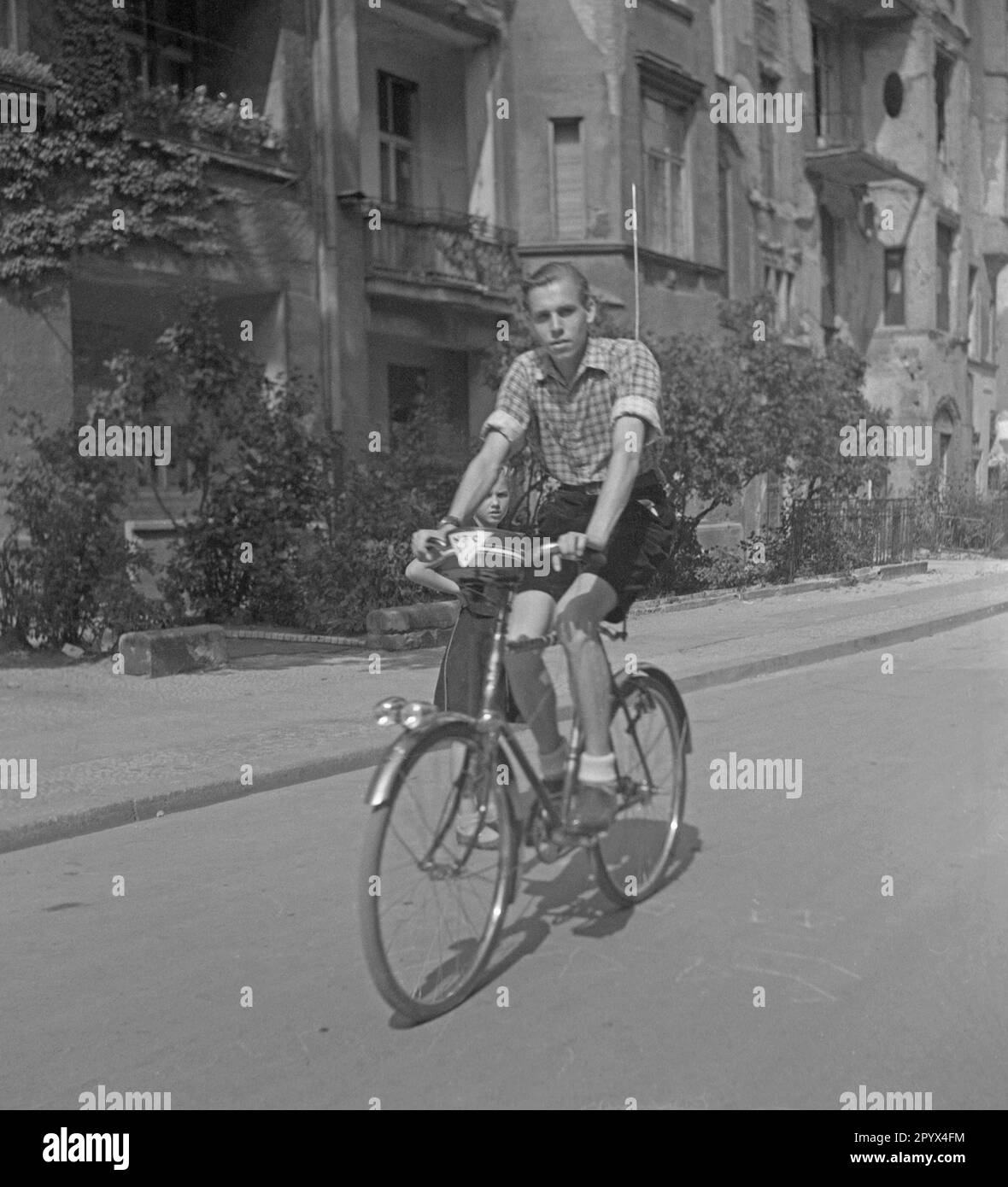 Undated photo of a cyclist in shorts and plaid shirt riding a men's bike equipped with an antenna and a transistor radio in West Berlin. On the bike, a sign of the American broadcaster National Broadcasting Company (NBC). Stock Photo