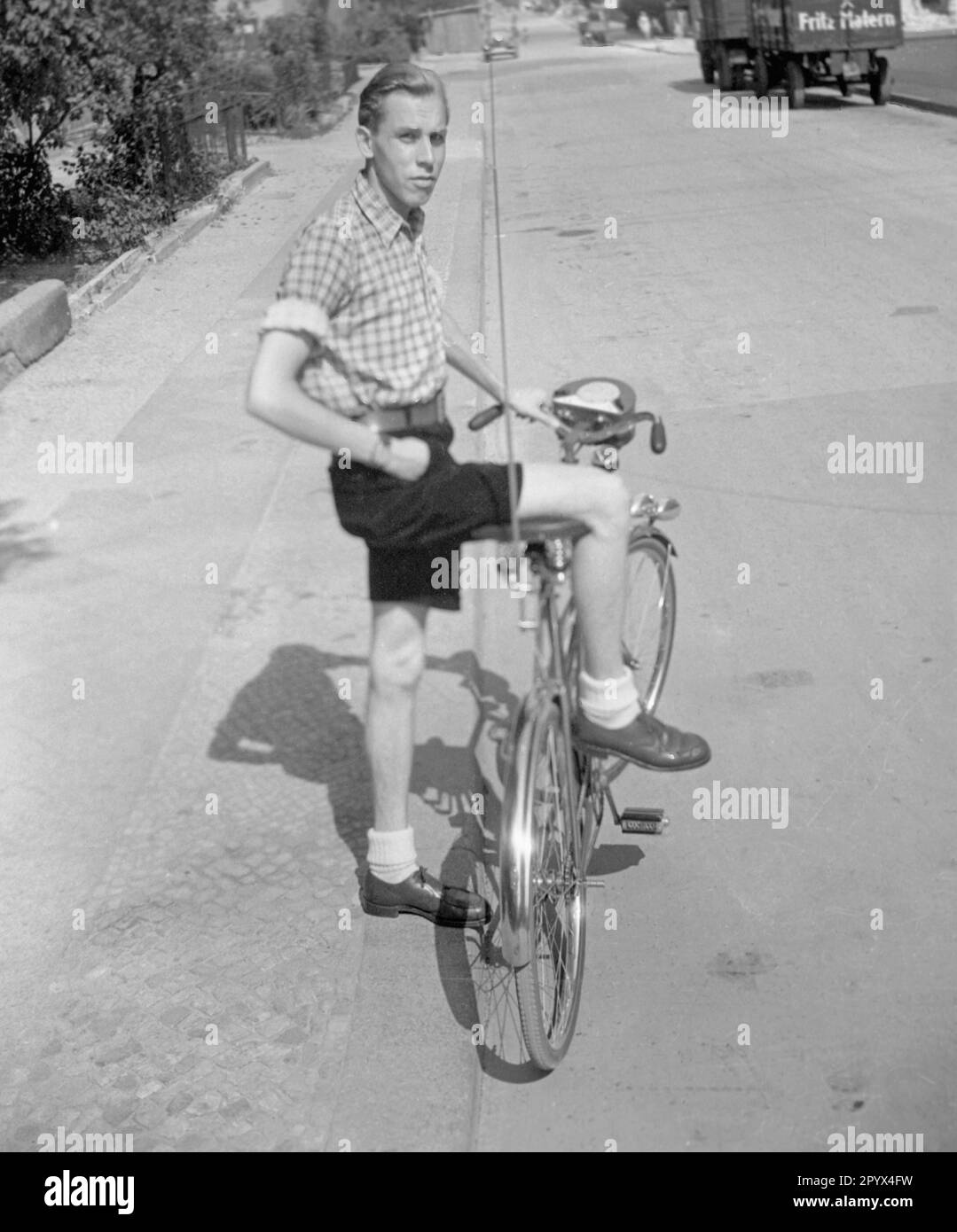 Undated photo of a cyclist in shorts and plaid shirt riding a men's bike equipped with an antenna and a transistor radio in West Berlin. In the background, a truck of the Fritz Matern Kohlenhandlungsgesellschaft (coal trading company) mbH in Berlin. Stock Photo