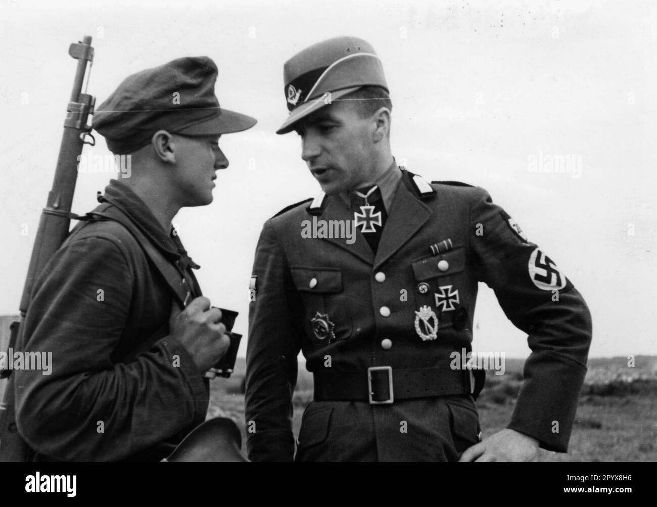 A highly decorated officer (Oberstfeldmeister / Hauptmann) of the Reich Labor Service talks to a member of the RAD assigned to guard duty during a visit to the Atlantic Wall. In addition to the Knight's Cross, he is decorated with the German Cross in Gold, the Infantry Storm Badge and the Iron Cross I. Class. Under the right pocket flap he wears the party badge of the NSDAP. Photo: Creten [automated translation] Stock Photo