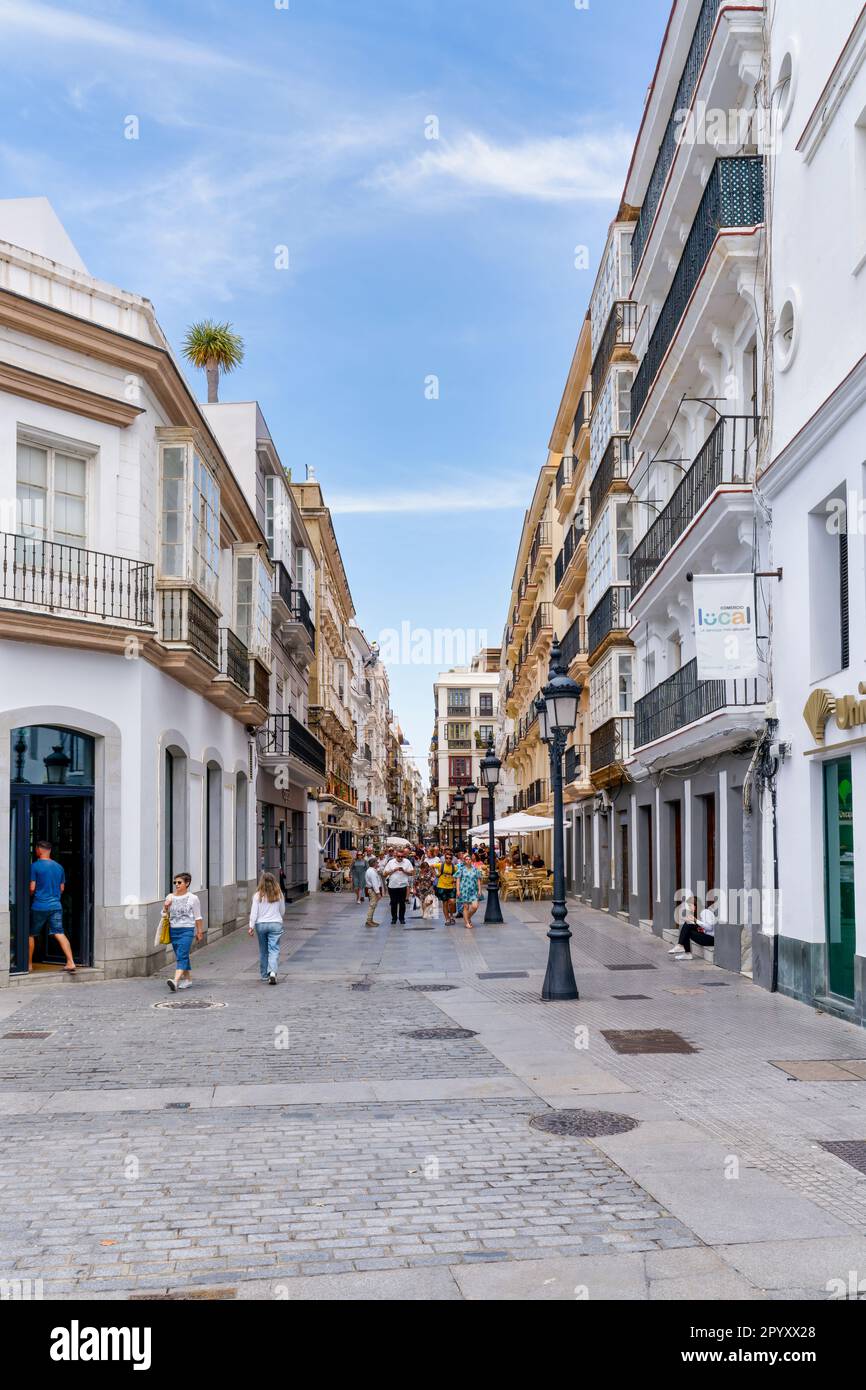 Tourists amble long a cobbled street, under a bright blue sky, in Cadix, Spain Stock Photo