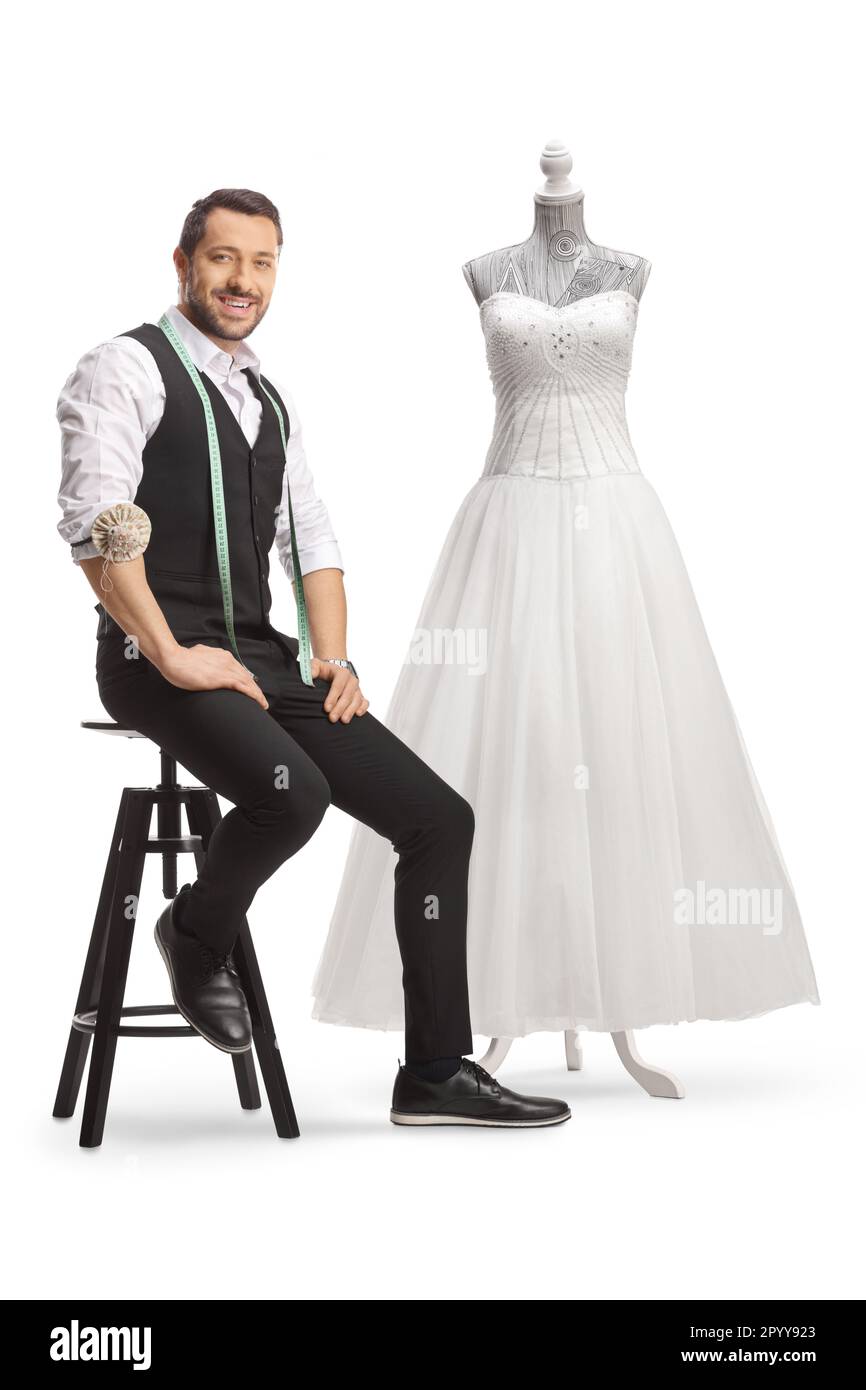Young tailor with a measuring tape smiling at camera and sitting next to a bridal gown isolated on white background Stock Photo
