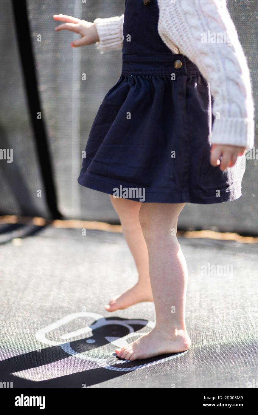 little barefoot girl in pinafore playing outside in sunshine on trampoline in the backyard Stock Photo