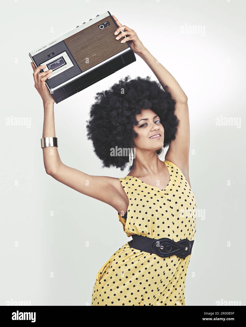 Moving to the groove. A young woman wearing a 70s retro jumpsuit holding a cassette player and striking a disco pose. Stock Photo