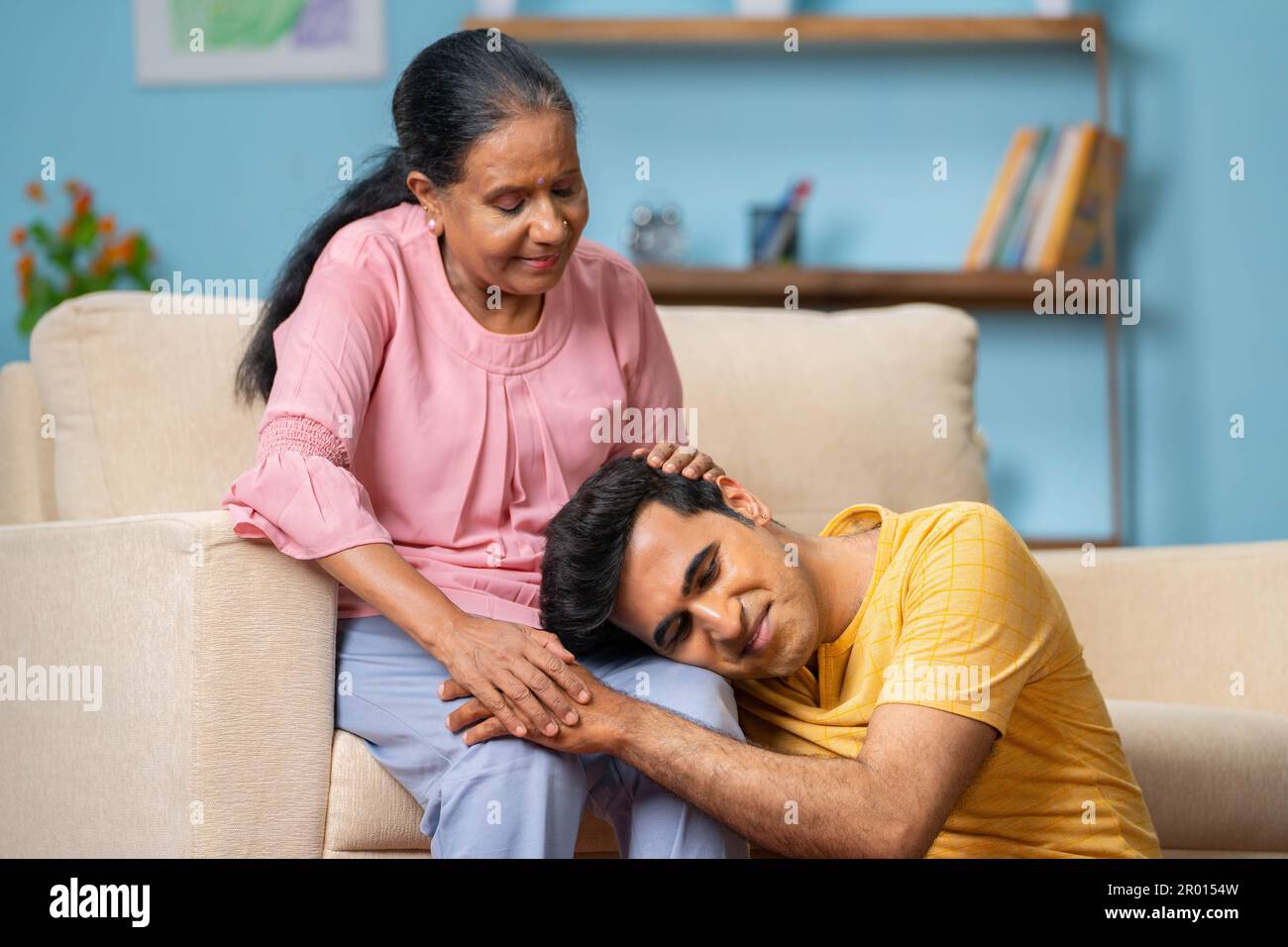 Indian senior mothers talking to adult son by consoling while son sleeping on mothers lap at home - concept of motherhood, parental support and Stock Photo