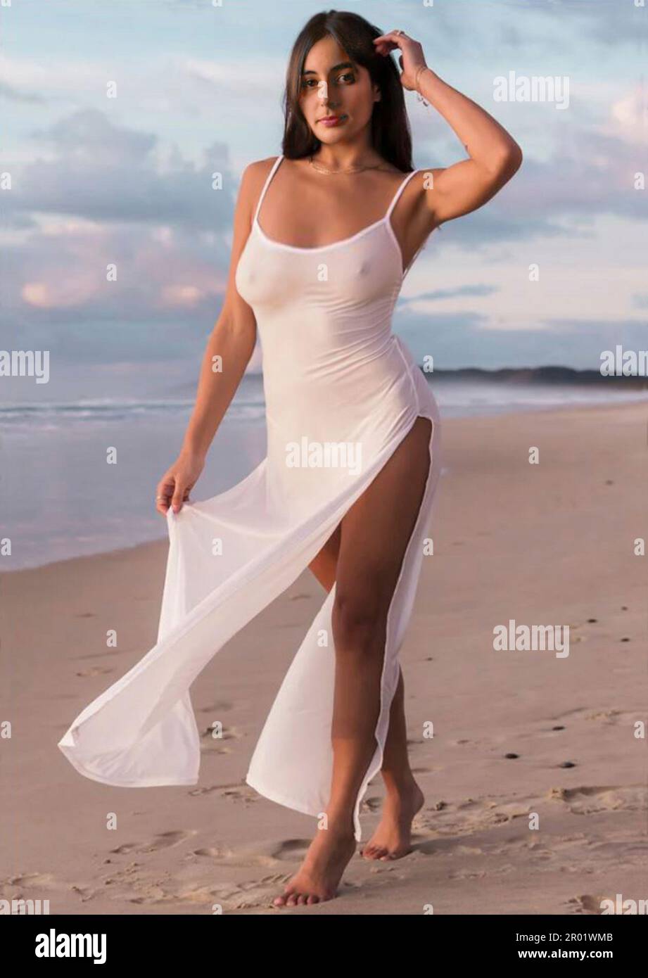 Glamorous brunette young woman wearing a long see-through white dress with high split side, walks along a deserted beach at sundown.  MODEL RELEASED Stock Photo