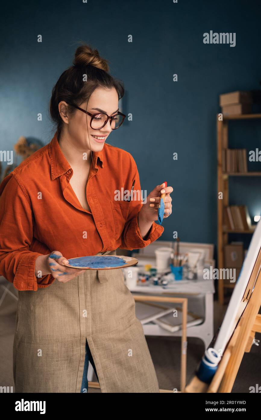 side view focused ethnic woman in casual clothes stands near the canvas holds a palette with paints in her hands looks at the canvas and thinks about Stock Photo