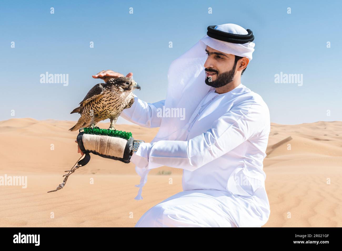 Middle-eastern man wearing traditional emirati arab kandura in the desert and holding a falcon bird - Arabian muslim adult person at the sand dunes in Stock Photo
