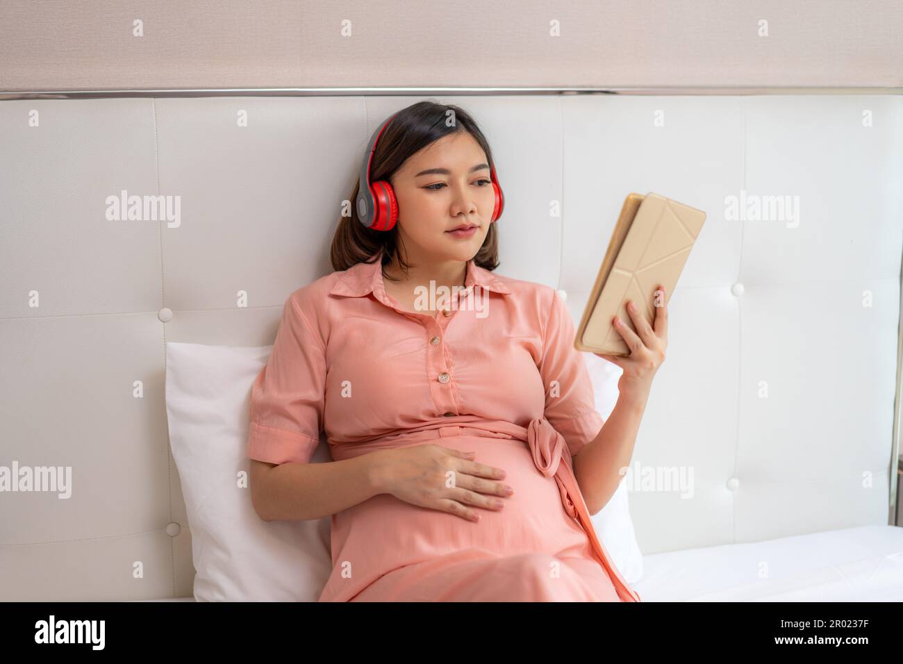 Happy young pregnant Asian woman wearing red headphone and sitting in her bed relaxing while looking at social media on her computer tablet Stock Photo