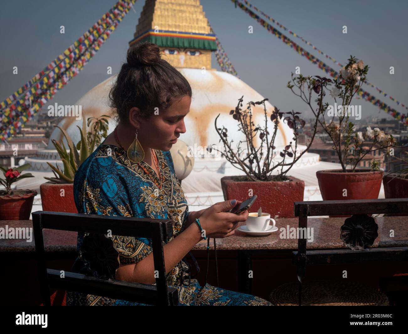 Stylish female backpacker hippie texting while having a cup of chai tea with Bouddhanath stupa (Nepal) in the background. Stock Photo