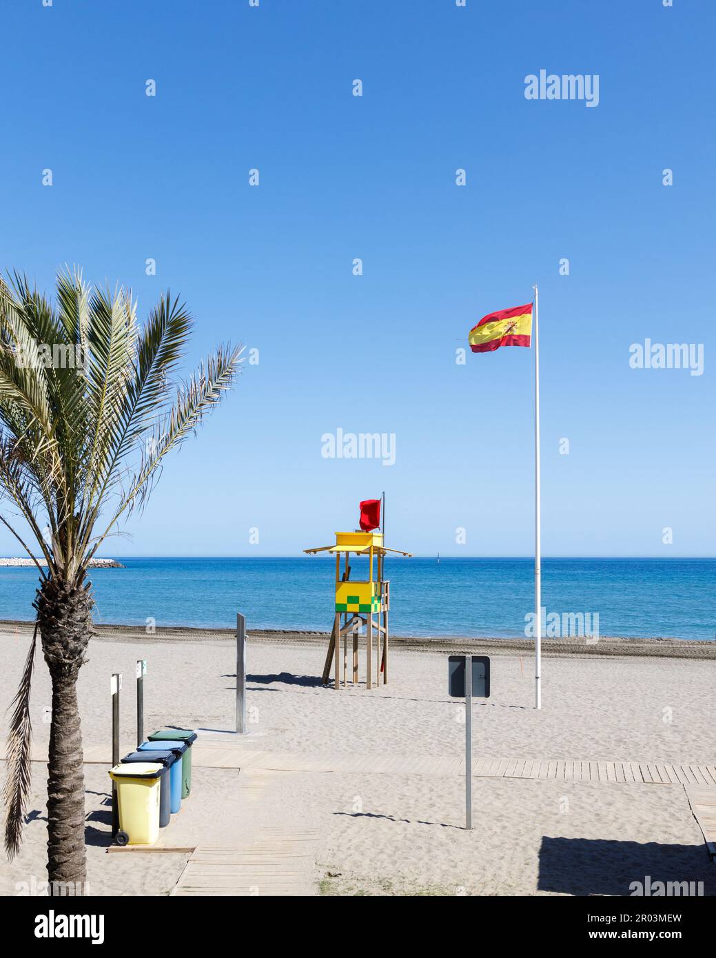 Beach with a palm tree in the foreground. Clear blue sky, deep sea, ocean, waving flag of Spain, sand, colored trash bins, rescue booth, summer Stock Photo