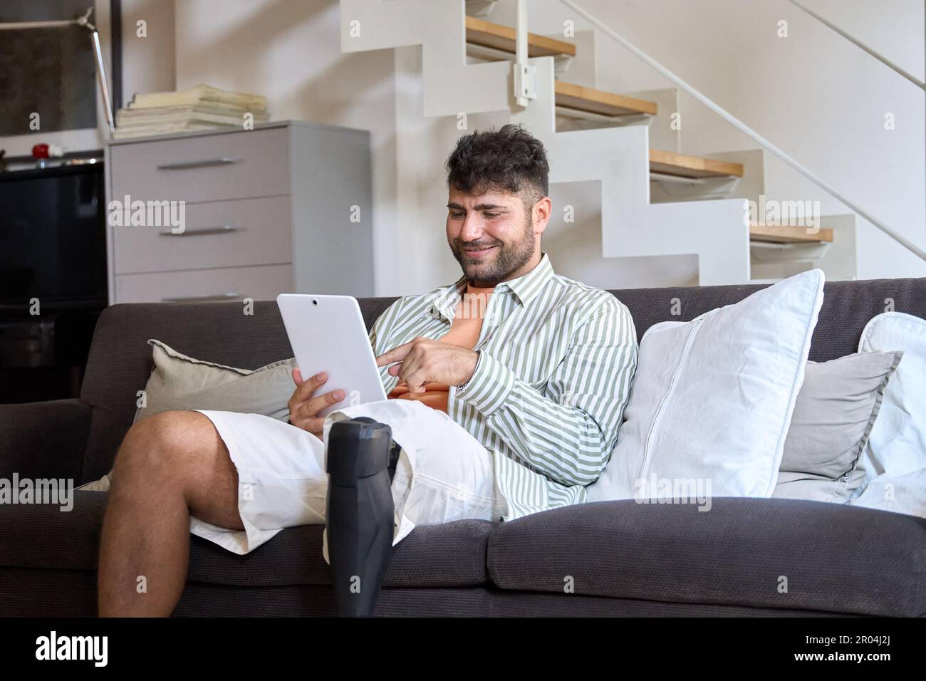 Happy man with disability leg prosthesis sitting on couch at home using tablet. Stock Photo