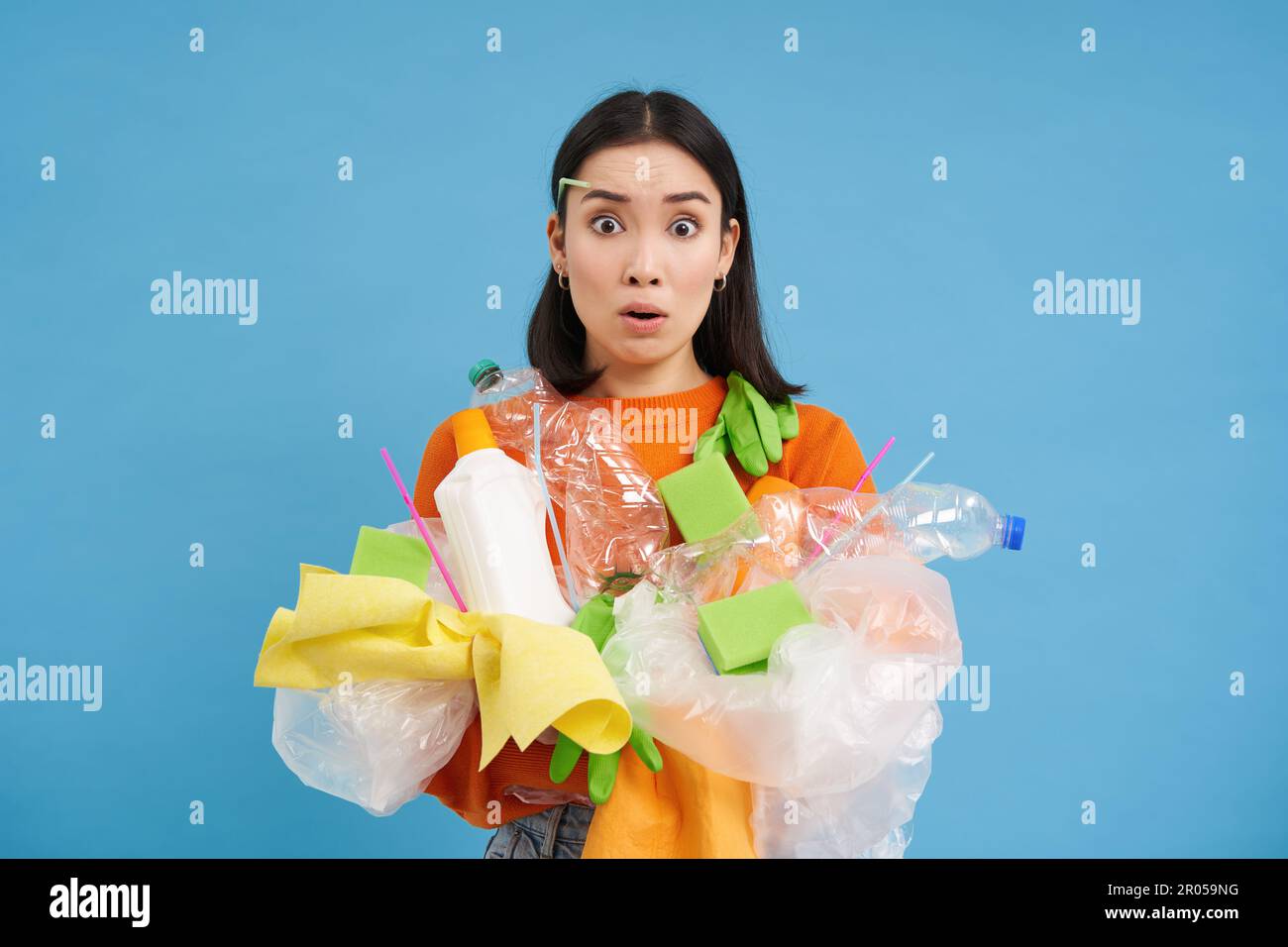 Young confused asian girl with different types of plastic, learns how to recycle, saving enviroment, sorting garbage, blue background. Stock Photo