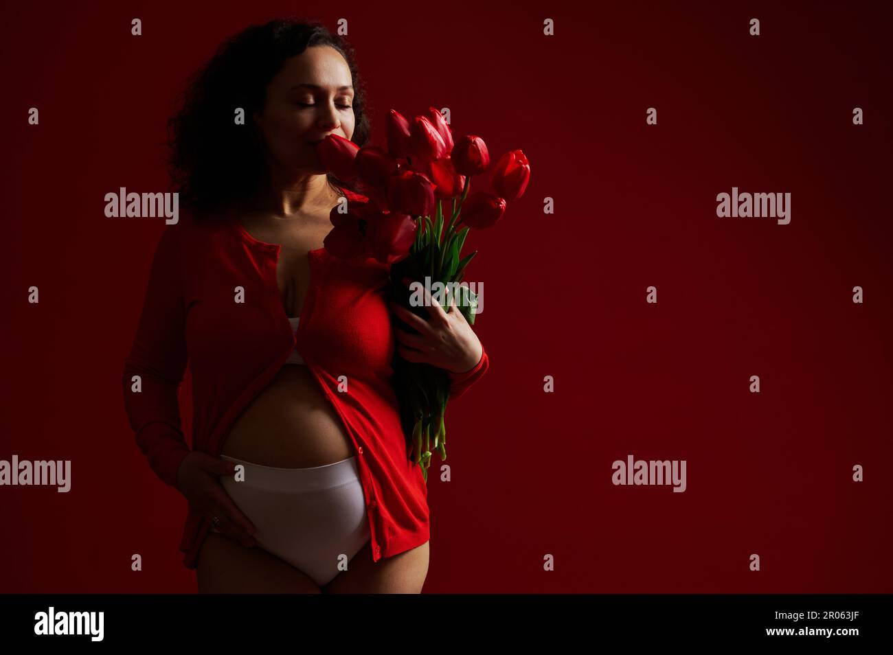 Gorgeous Latin American adult pregnant woman caressing her belly, sniffing a cute bouquet of red tulips for Mother's Day Stock Photo