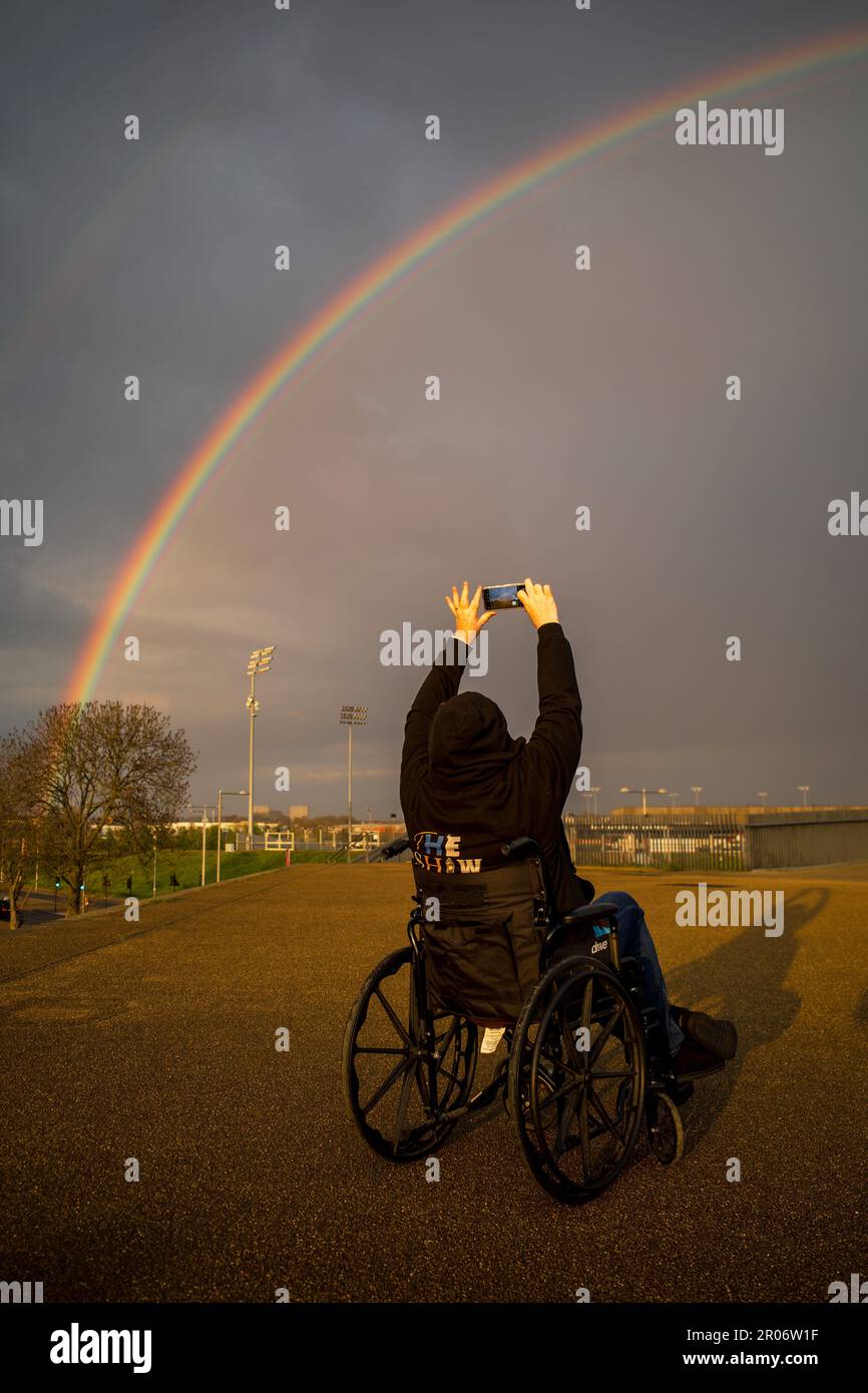 A woman wearing a black hoodie and sat in a wheelchair takes a photograph of a rainbow using her mobile phone. Location is the Olympic Park, Stratford Stock Photo