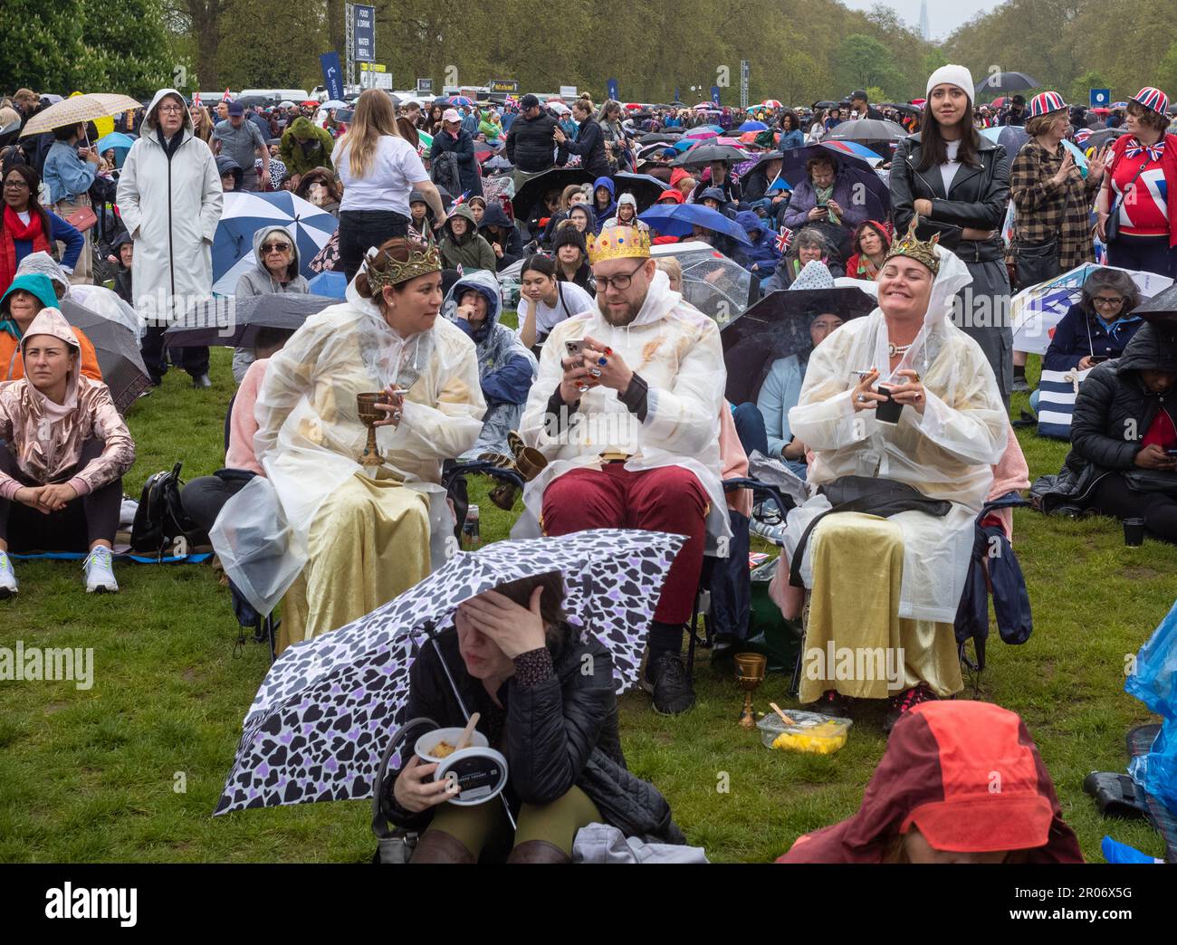 Three people wearing fake crowns and raincoats smoke cigarettes and drink from goblets in Hyde Park, London, UK. In steady rain, a sea of patriotic Br Stock Photo