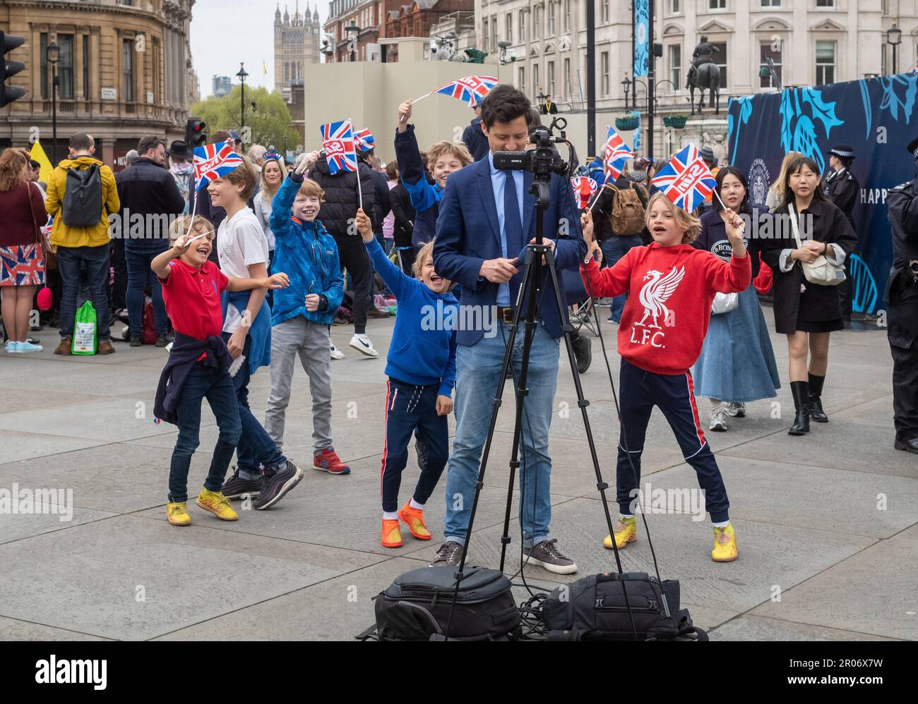 In the heart of London's Trafalgar Square, a news television reporter stands in front of his camera. Behind him, a group of young children dance and w Stock Photo