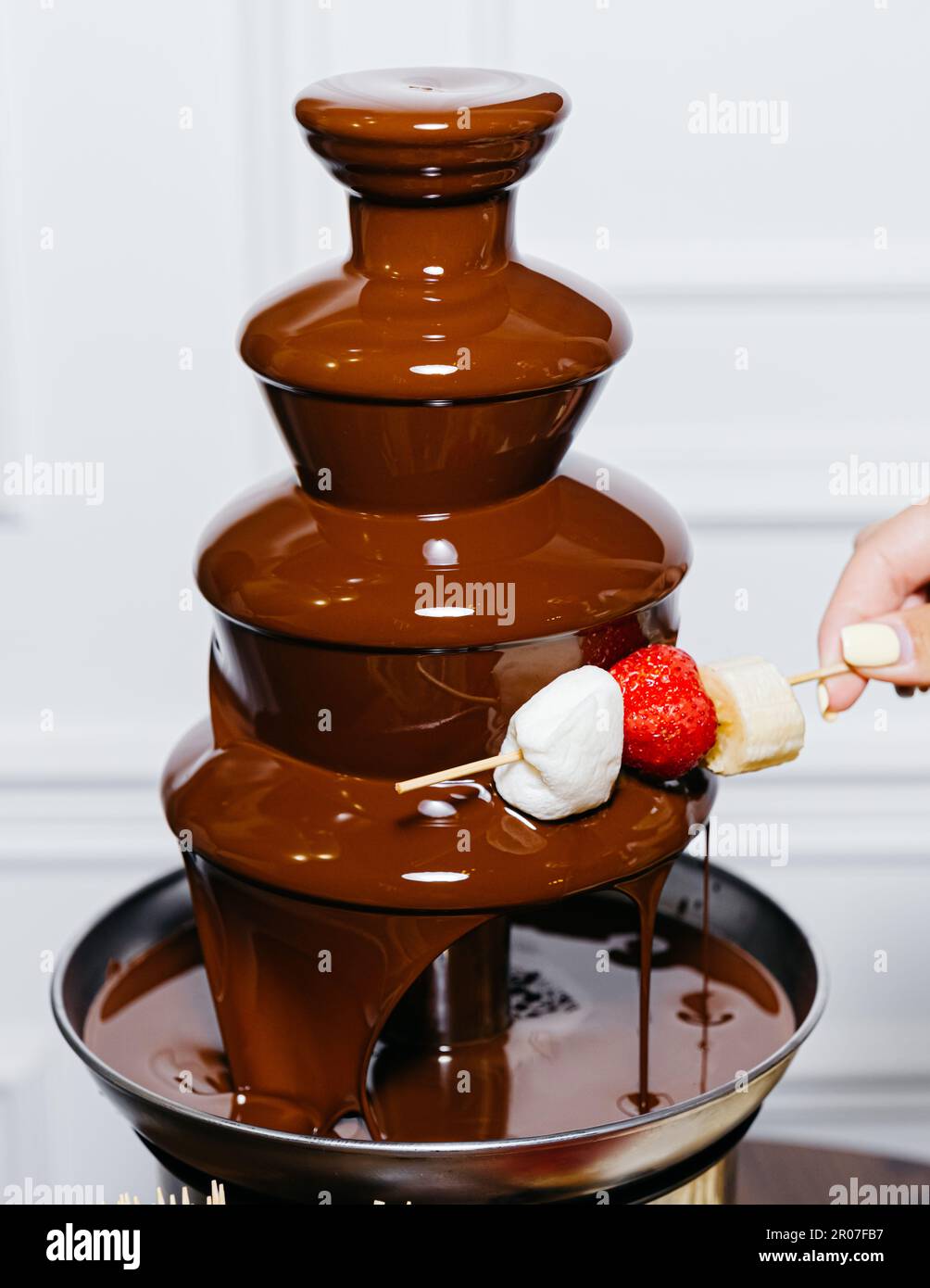 A person is enjoying a sweet dessert of banana, marshmallow, and strawberry pieces drizzled in chocolate from a chocolate fountain. Stock Photo